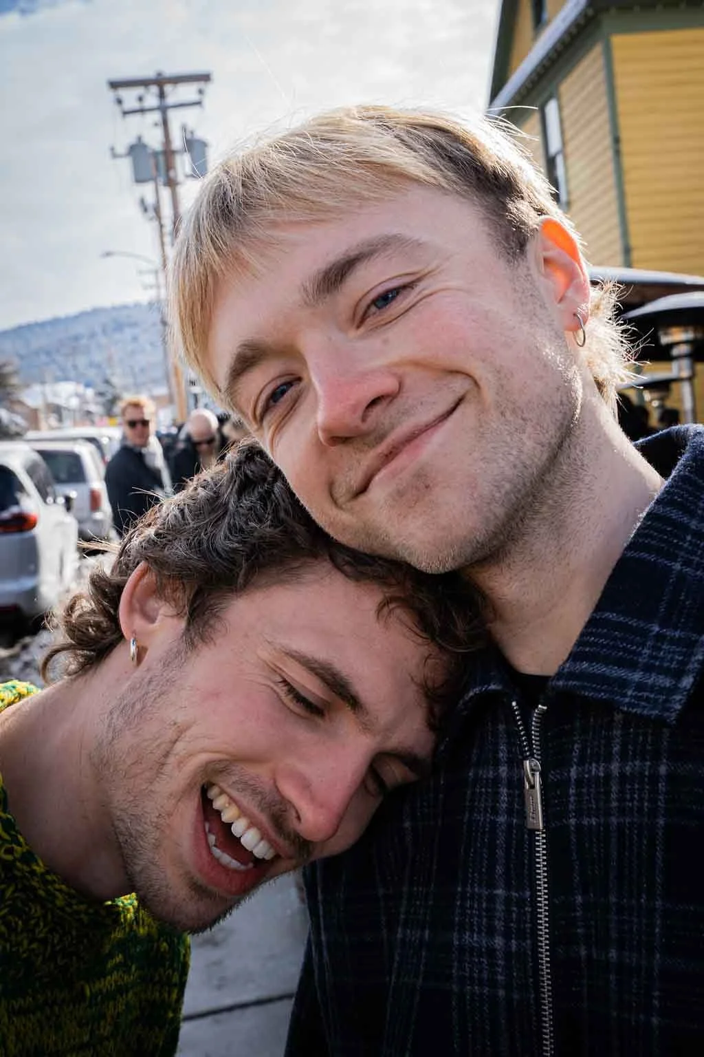 Two young men sharing a hug; the man on the left has curly hair and is wearing a colorful sweater, smiling with closed eyes, and the man on the right has straight blonde hair and is wearing a dark checkered jacket, smiling gently. They are outdoors w