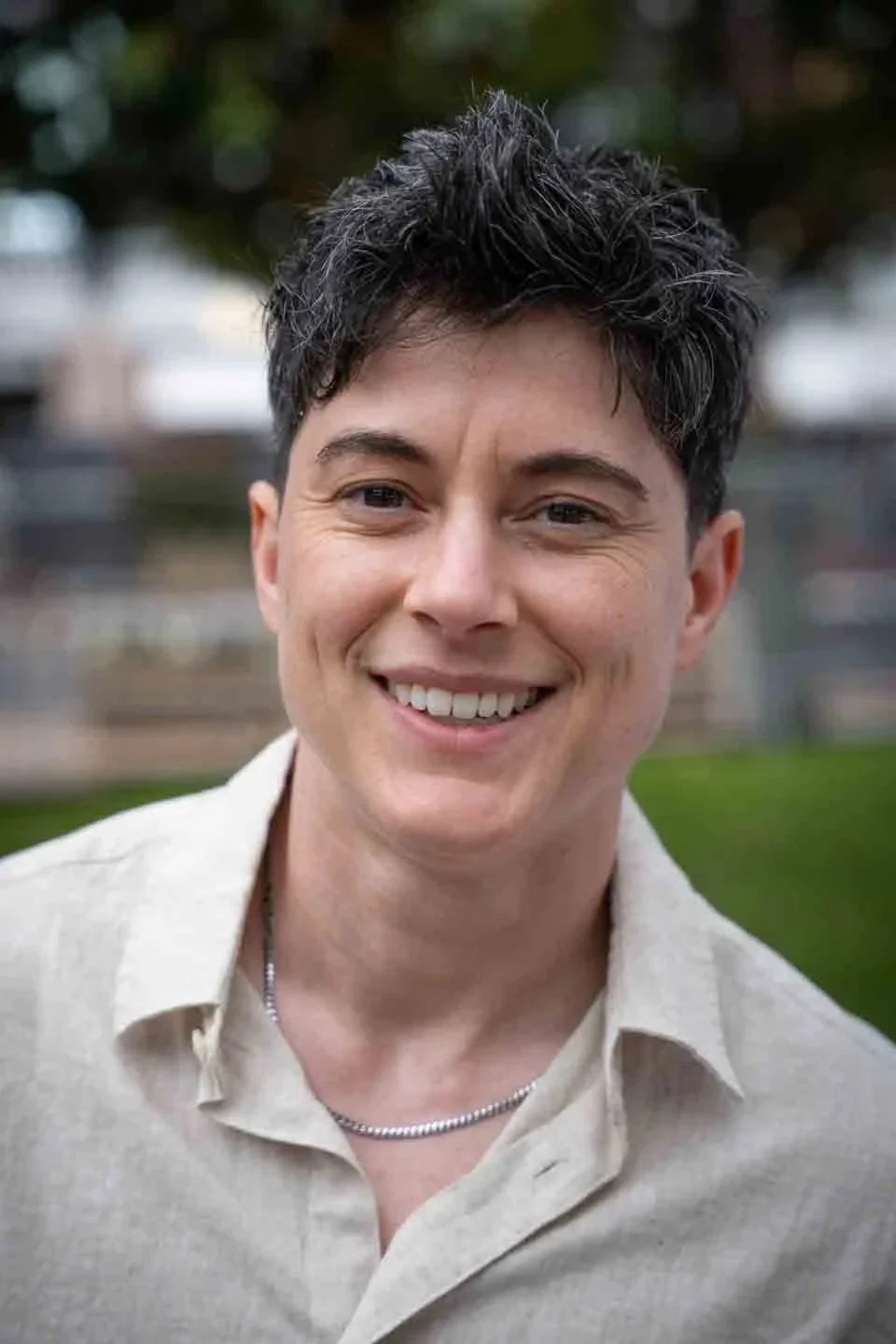 A smiling woman with short, dark, wavy hair, wearing a beige shirt and a silver chain necklace, outdoors with blurred trees and grass in the background.