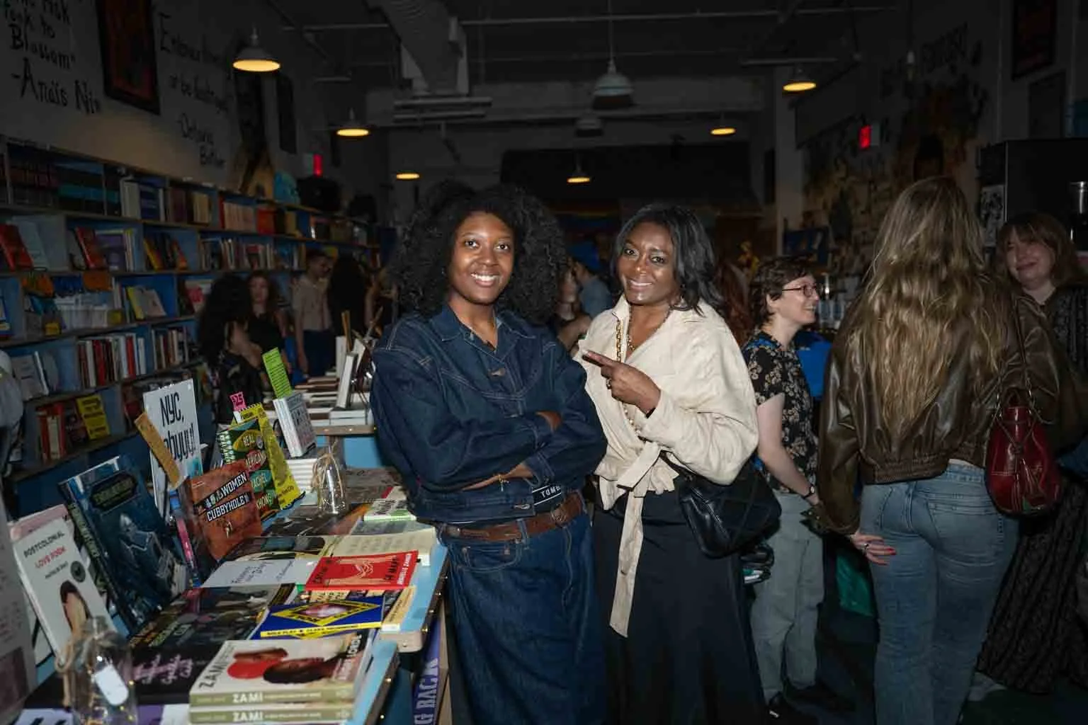 Two women smiling and posing for the camera at a book signing event, with books and shelves behind them and other attendees in the background.