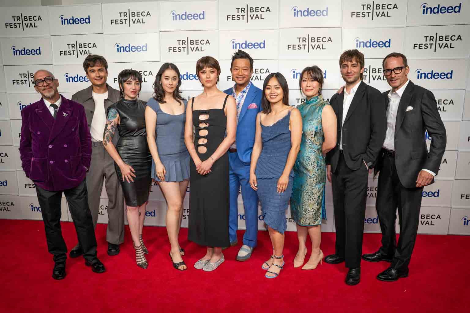 A group of ten people standing on a red carpet at the Tribeca Festival, posing for photos in front of a backdrop with Tribeca Festival and Indeed logos. They are dressed in formal to semi-formal attire, smiling, and standing close together.