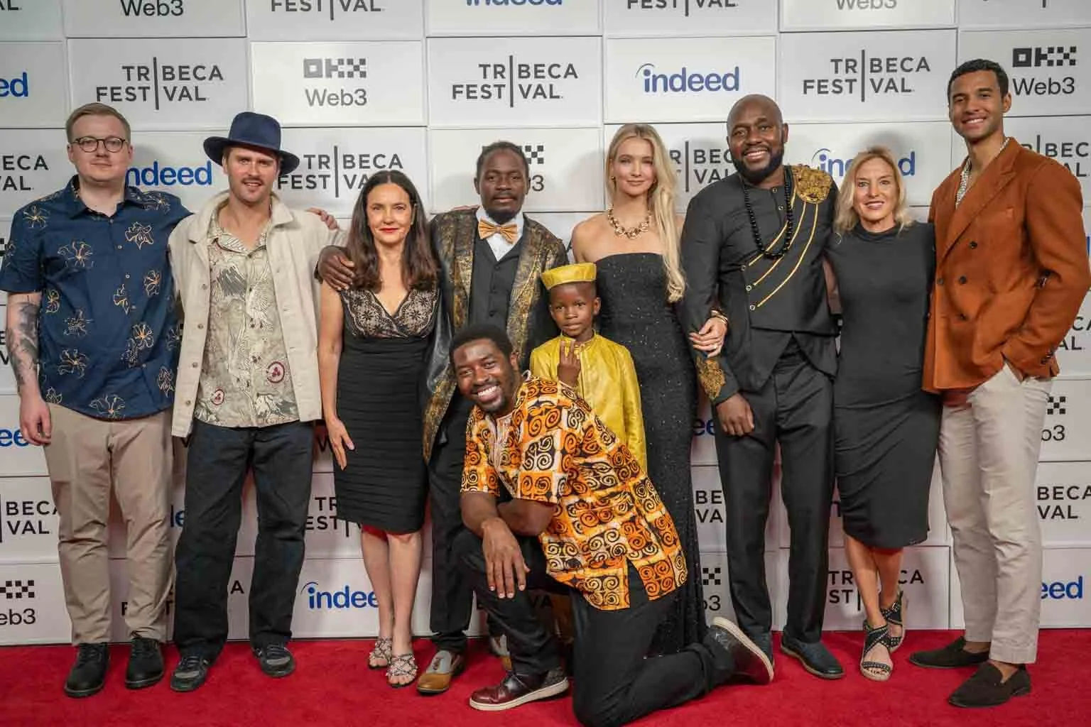A group of ten diverse individuals posing together at the Tribeca Film Festival, standing in front of a white backdrop with logo posters including Tribeca, Web3, Indeed, and others. Some are smiling, dressed in various stylish outfits, with two kneel