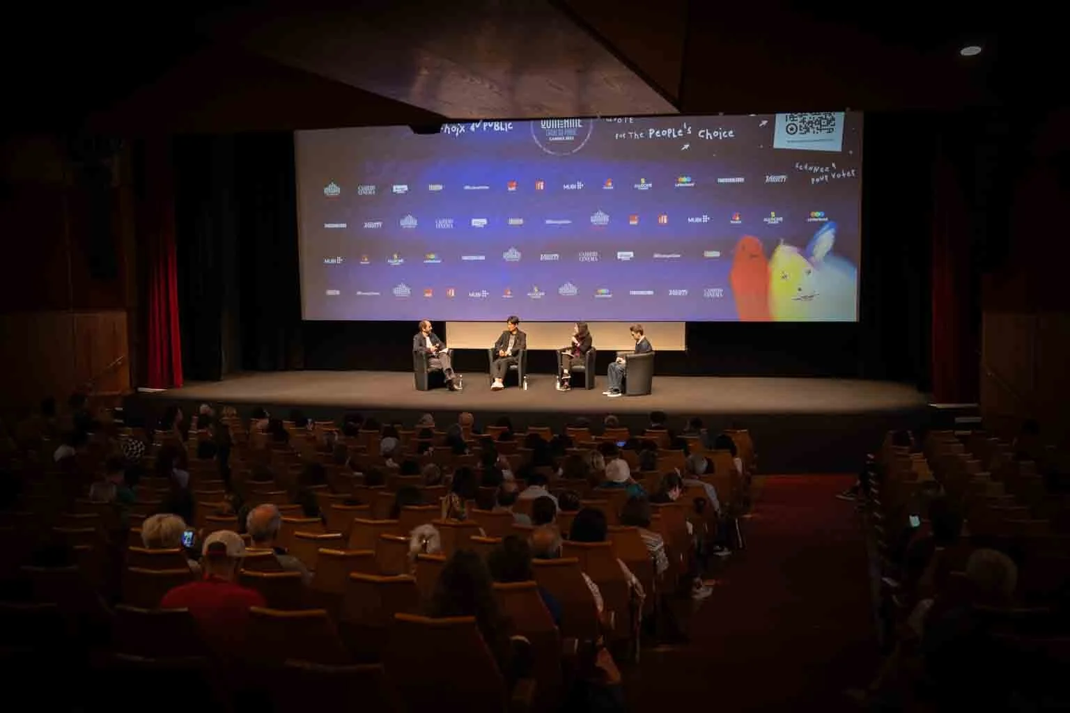 An auditorium with a stage where four people are seated and participating in a panel discussion. Behind them is a large screen displaying various logos and images.