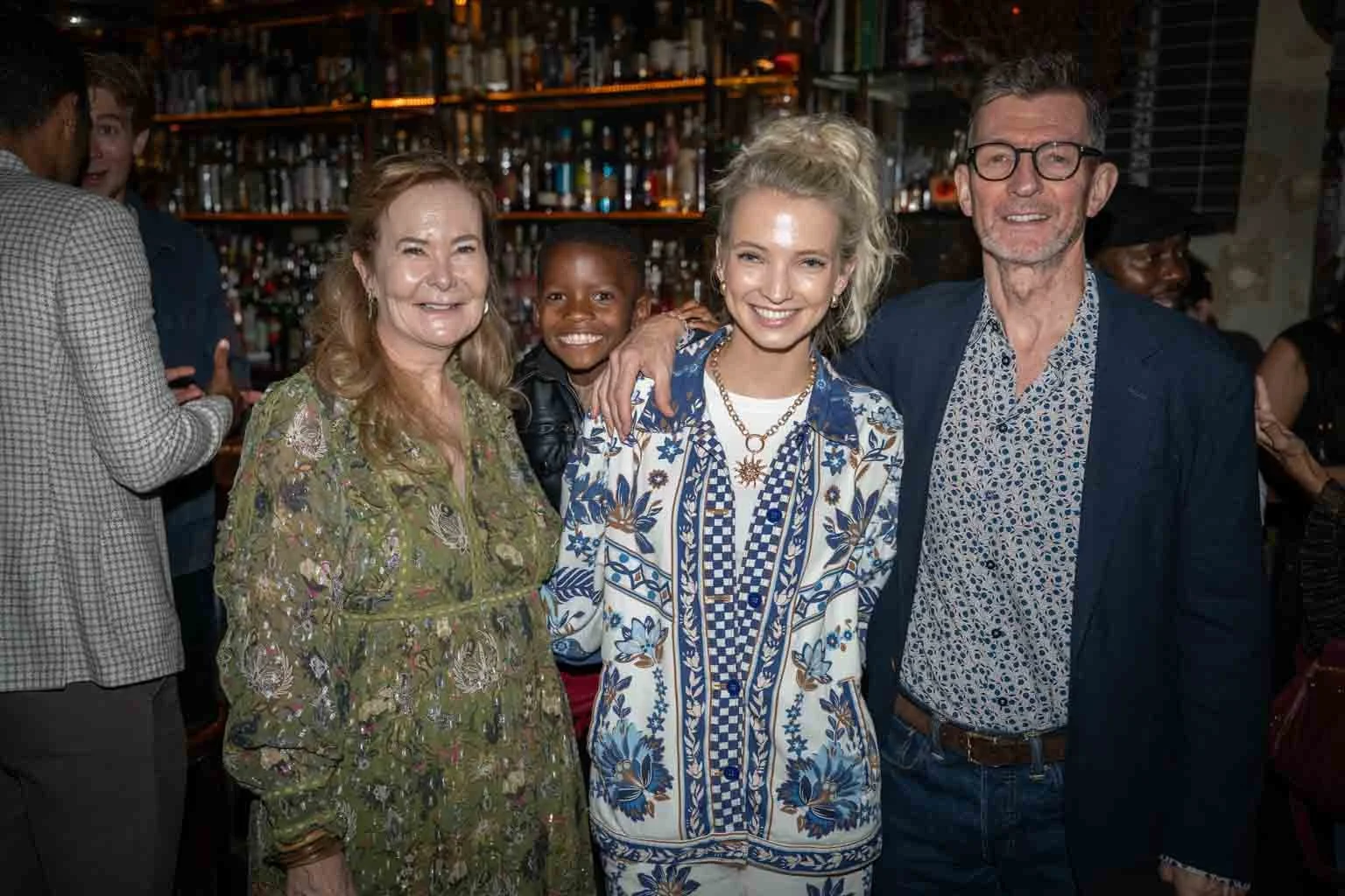 Group of four people smiling at a social gathering, with shelves of bottles behind them and others in the background.