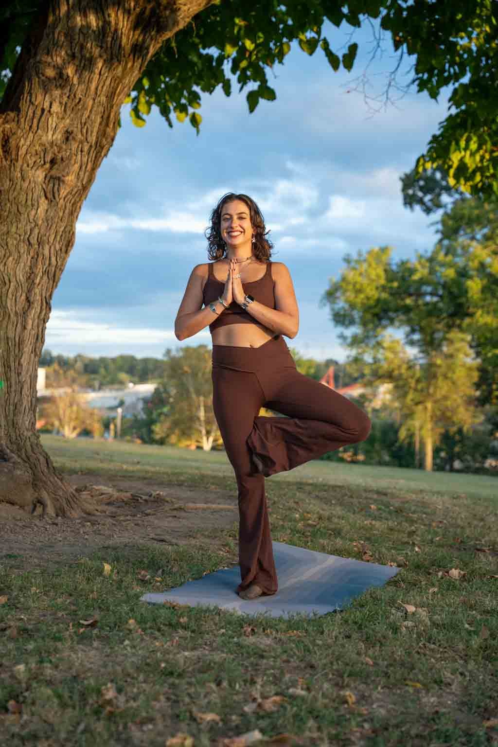 Sammie Gaynor - A woman practicing yoga outdoors under a large tree during the daytime, standing on a yoga mat, balancing in a tree pose with hands together in a prayer position.