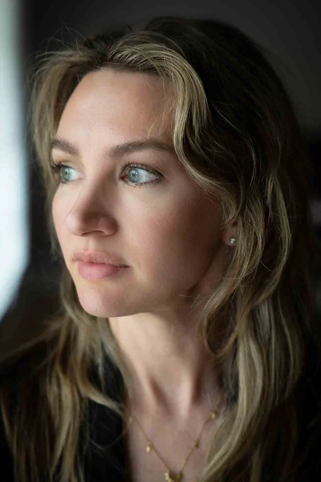 Minette Martin - Close-up portrait of a woman with long wavy hair and blue eyes, wearing a gold necklace, looking directly at the camera.
