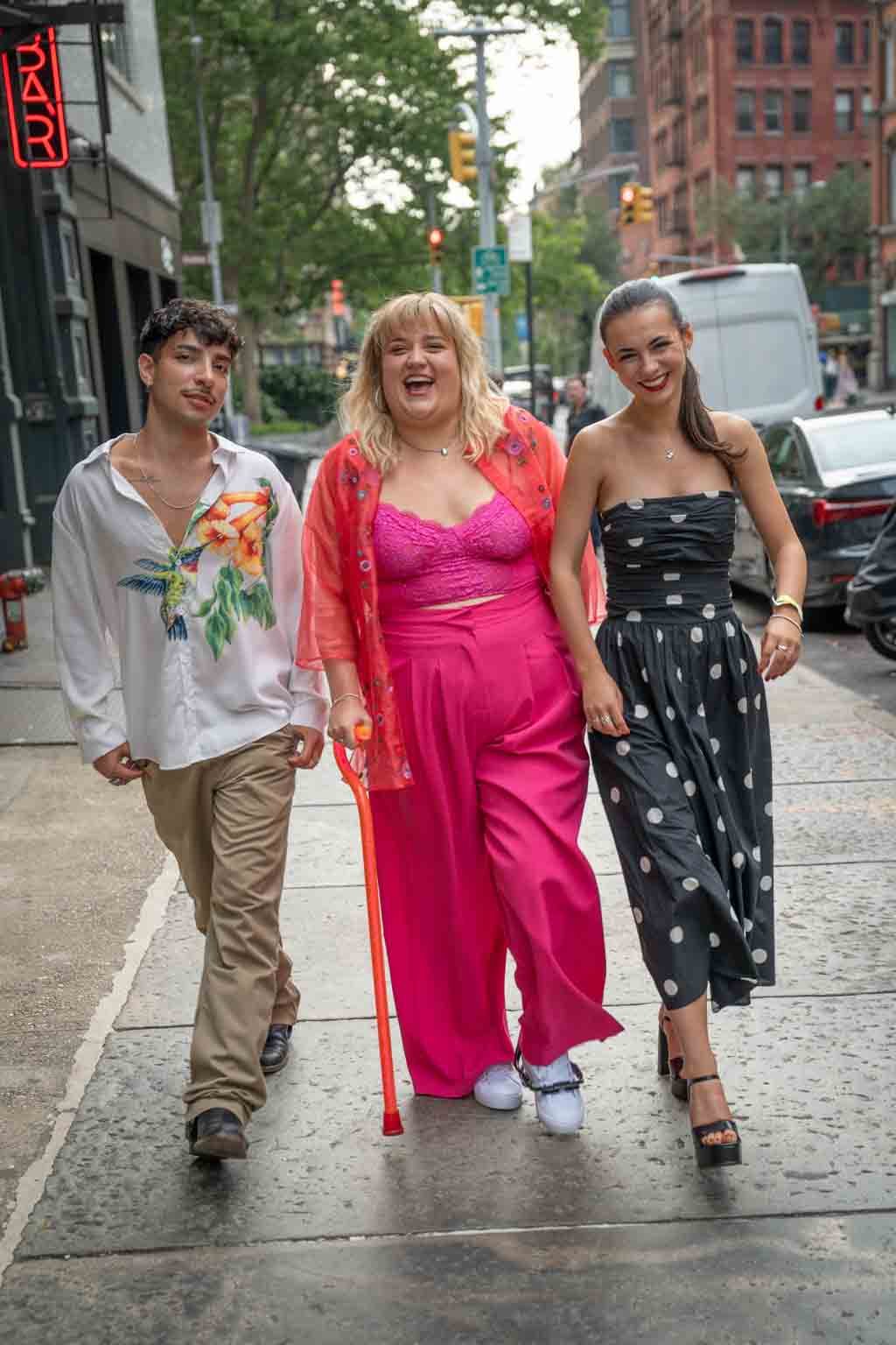 Three young women walking together on a city sidewalk, smiling and dressed in colorful and stylish clothing, with cars parked along the street behind them.