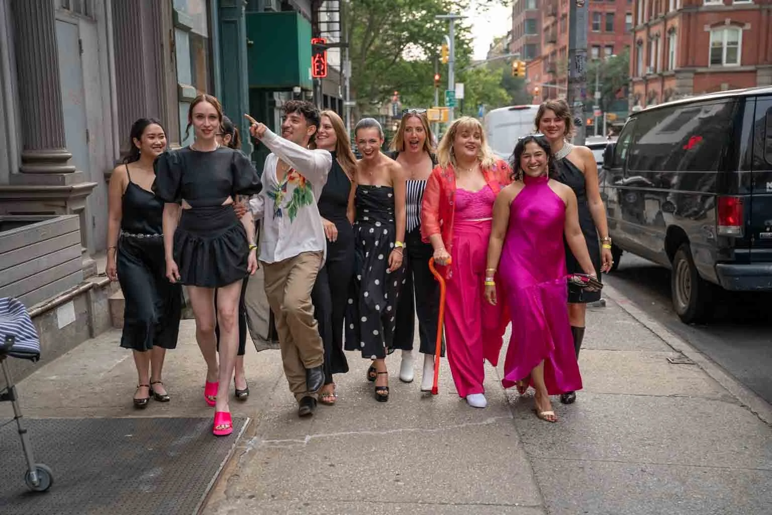 Group of fashionable women walking on city sidewalk, dressed in stylish outfits, some smiling, as traffic lights and vehicles are visible in the background.