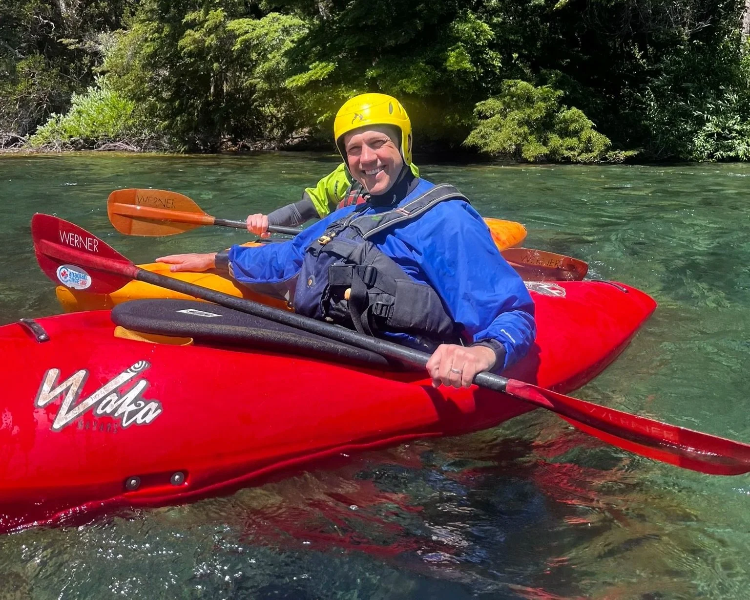 Coach Joe Miller grins from inside a bright red single-person kayak. He is wearing a neon yellow helmet, bright blue jacket, and safety gear, and he's holding a black and red paddle across the boat. A clear green creek and trees surround him.
