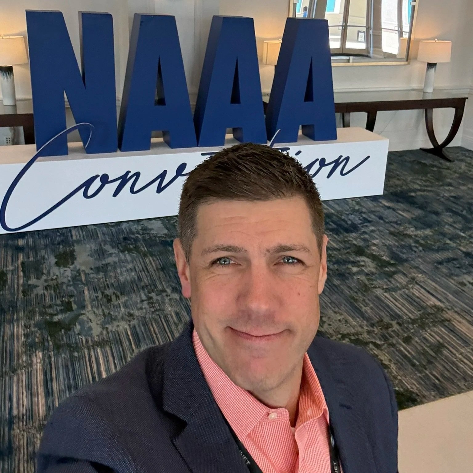 Coach Joe Miller smiles up at the camera. He's wearing a navy suit and salmon dress shirt. Behind him is a big custom display set up in a hotel lobby that announces 'NAAA Convention' in navy and white.