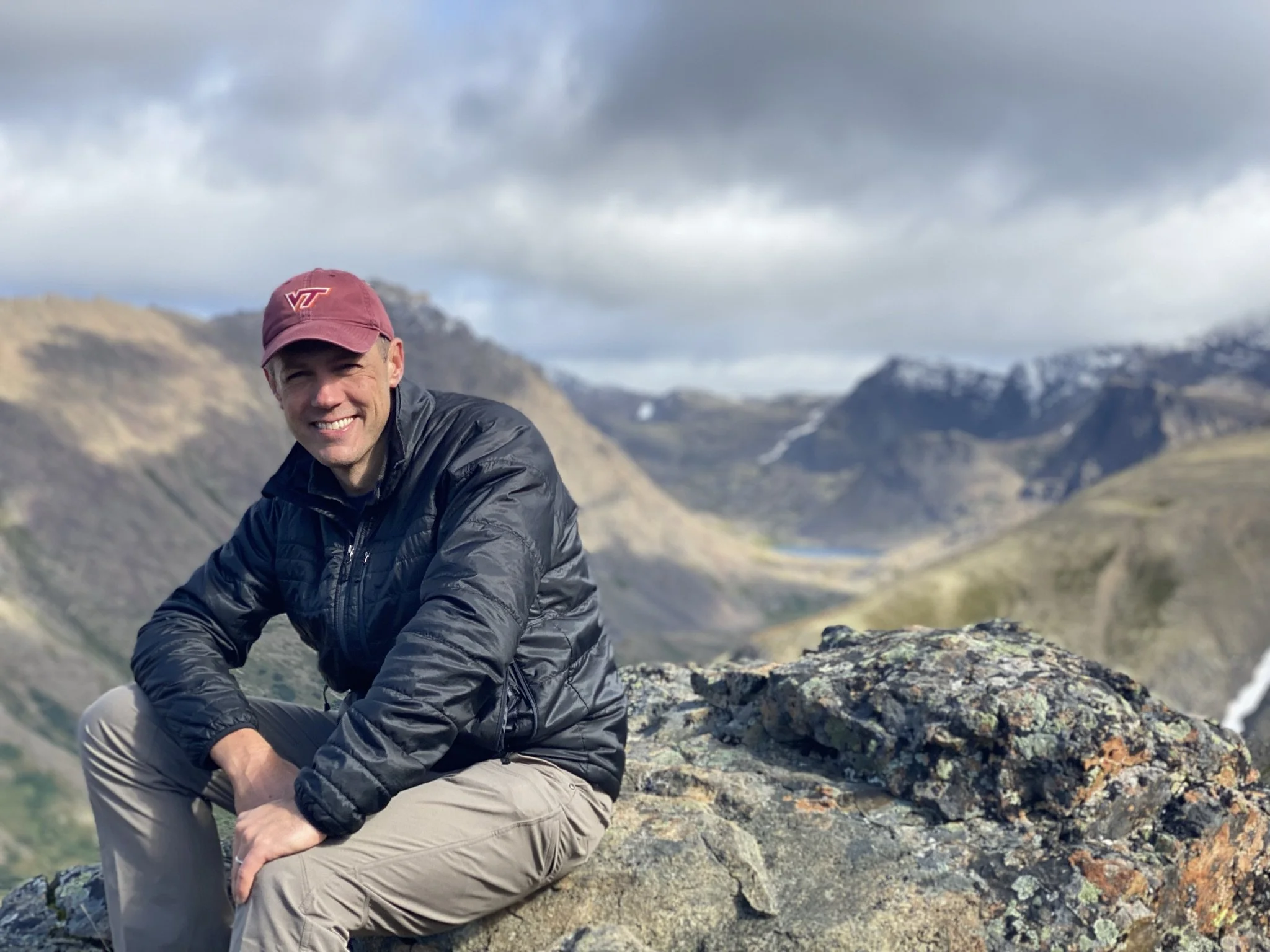 Coach Joe Miller perches on a rock at the top of a mountain. He is wearing a faded burgundy Virginia Tech ballcap, a shiny black thin puffer coat, and khaki athletic pants. Mountains and clouds and valleys are spread out behind him.