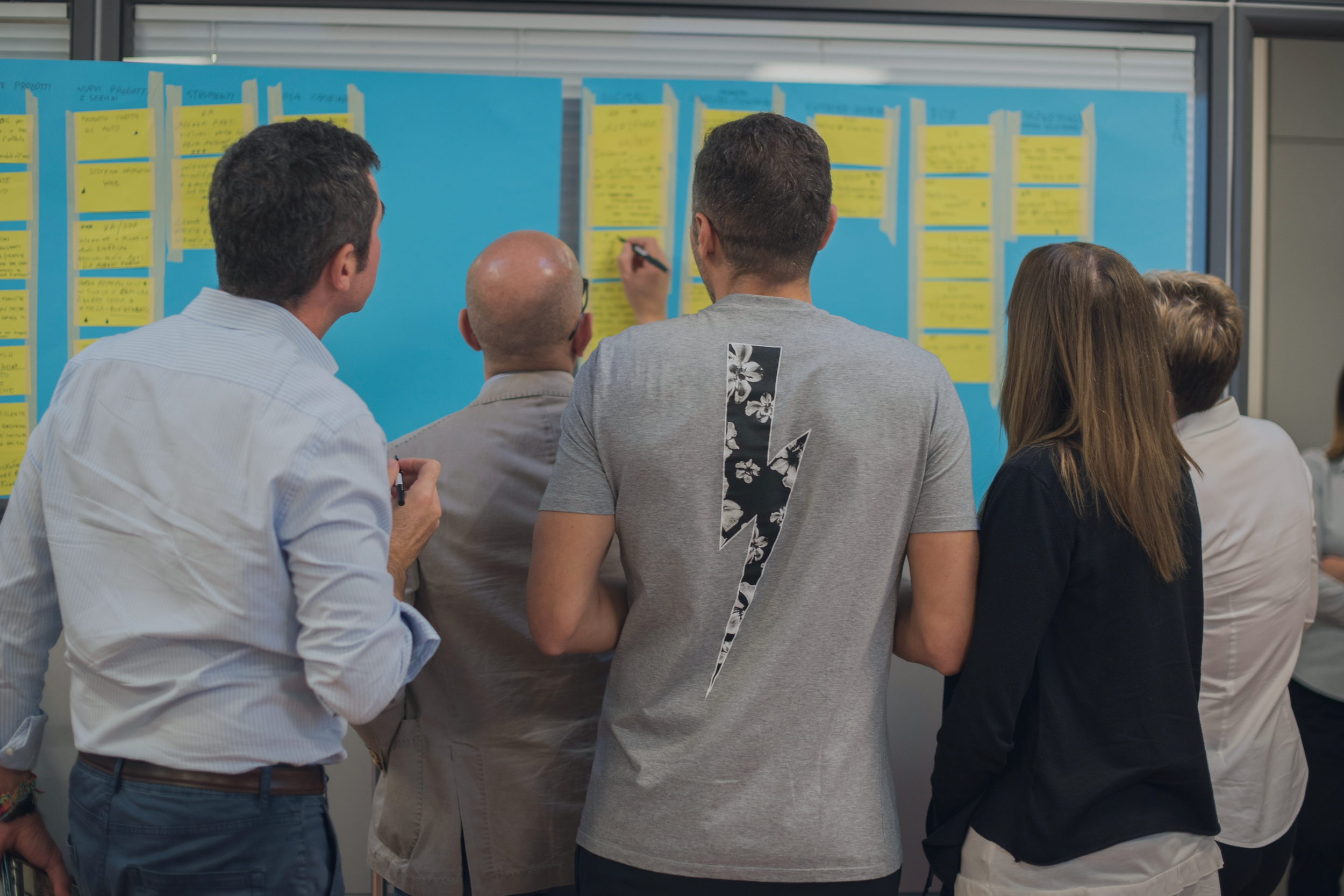 Five people crowd around a blue board covered in bright yellow index cards. They are wearing business casual clothes and seem to be working together to write on the cards.