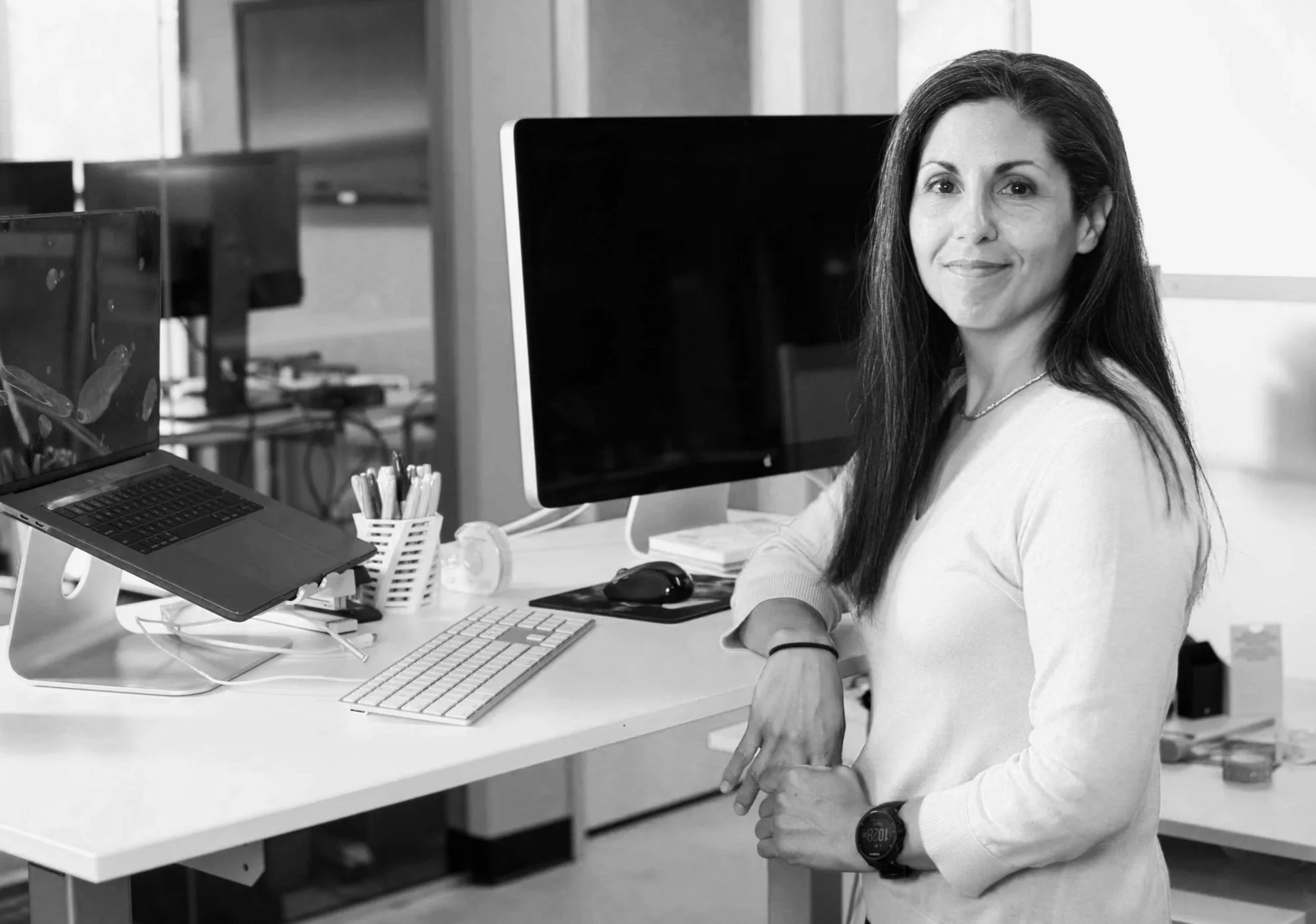 A woman with long, dark hair leans one arm on a standing desk and looks over her shoulder at the camera. She is wearing a white sweater and chunky athletic watch. Her desk has a large monitor, laptop, and keyboard on it.