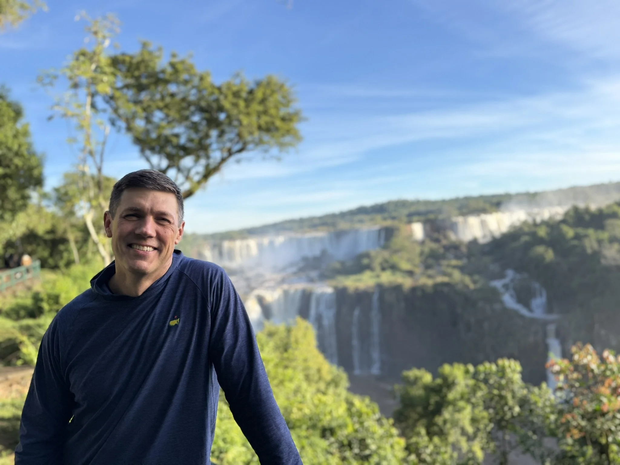 Coach Joe Miller leans on one arm and smiles at the camera - a beautiful, wide waterfall is visible behind him. Joe is wearing a dark blue pullover. The sun is lighting up the trees in the background.