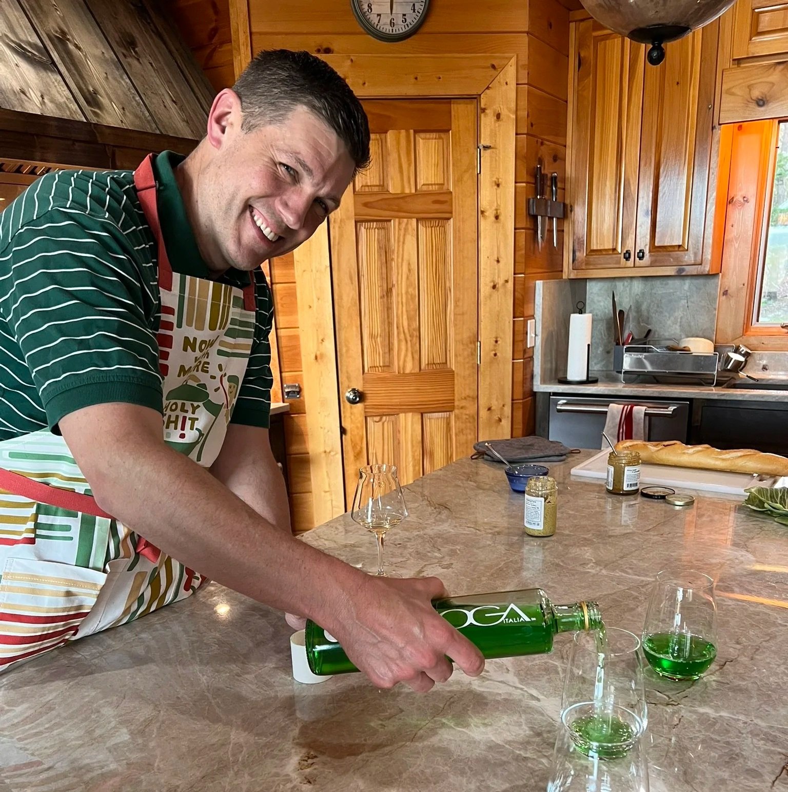 Coach Joe Miller stands at a granite countertop in a rustic, wood-paneled kitchen. He is wearing a holiday apron and grinning at the camera while pouring a vivid green beverage into glasses.