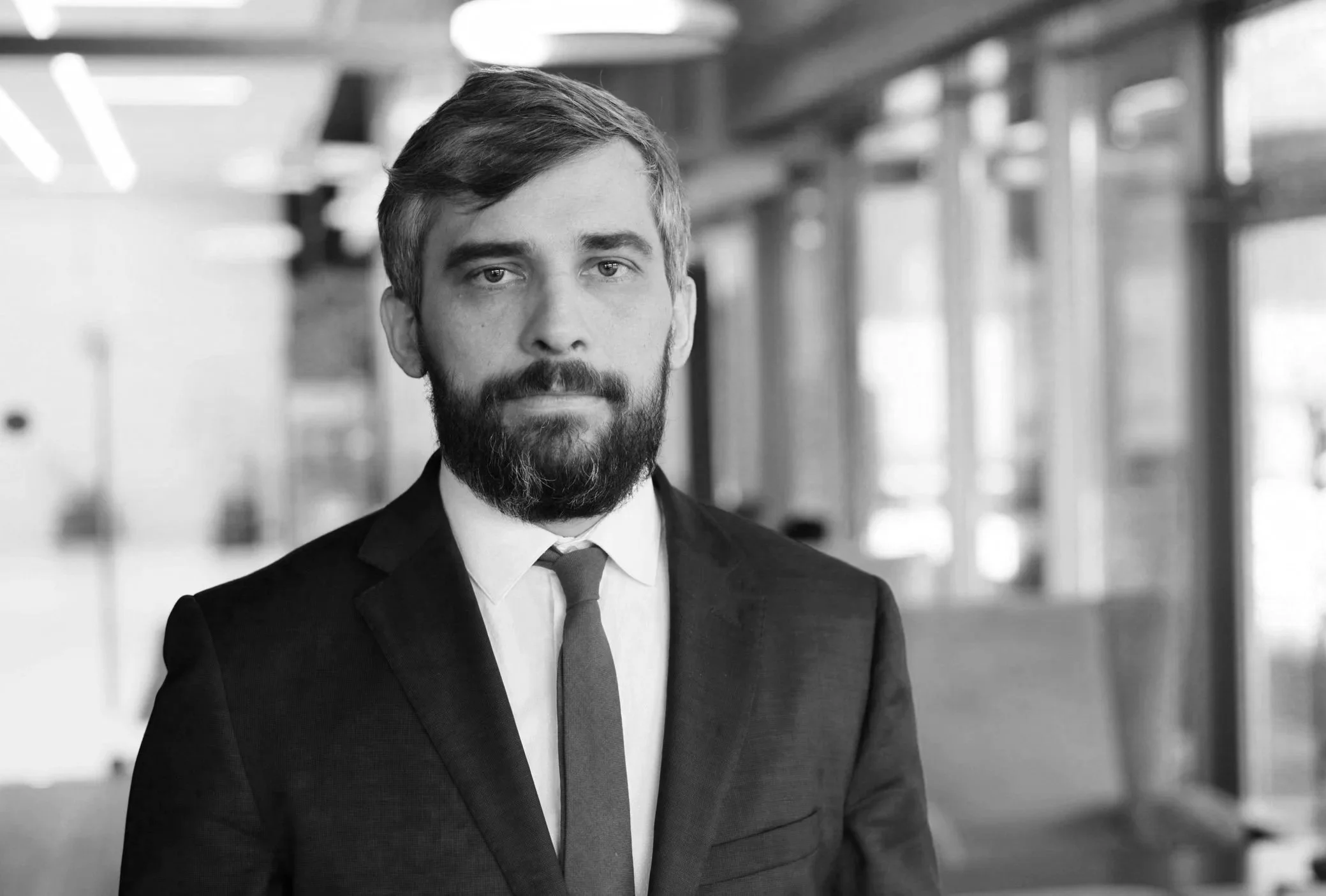 A man with salt-and-pepper hair and a dark beard looks seriously into the camera. He is wearing a dark business suit and patterned tie. He appears to be standing in an office hallway with fluorescent overhead lights and glass walls.