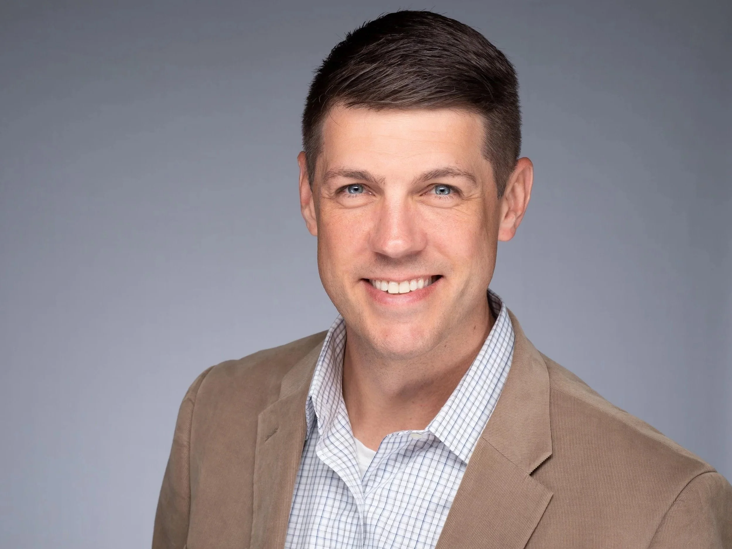 Professional headshot of Coach Joe Miller. Joe smiles openly at the camera - he has short dark-blond hair and striking blue eyes. He's wearing a tan sport coat and a blue checked shirt. The background is hazy blue-gray of a photo studio.