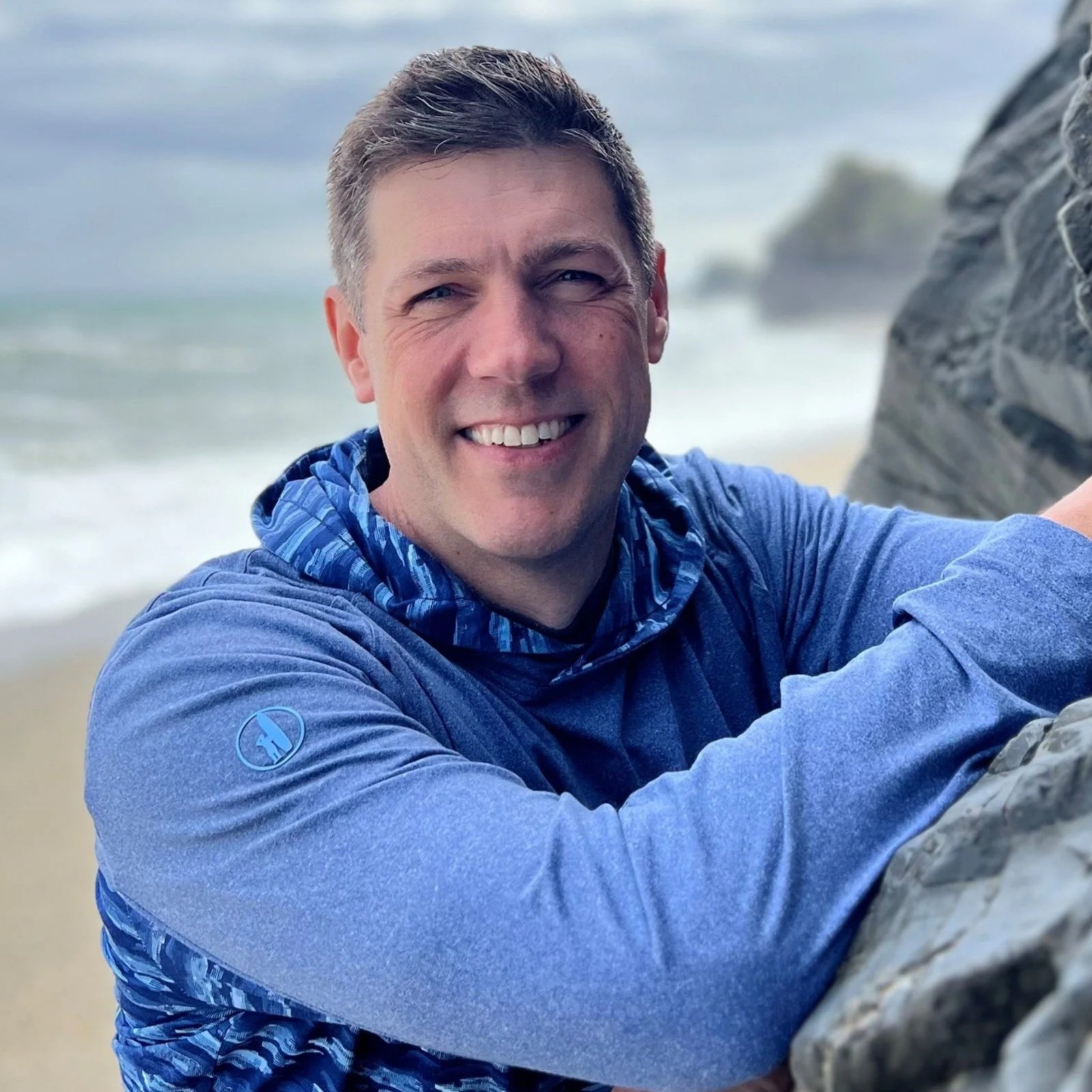 Coach Joe Miller smiles at the camera; he has short, spiky blond hair and blue eyes. He's wearing a light blue hoodie with a darker blue pattern lining the hood. His arms are propped on a large rock, a misty day at the ocean is visible behind him.
