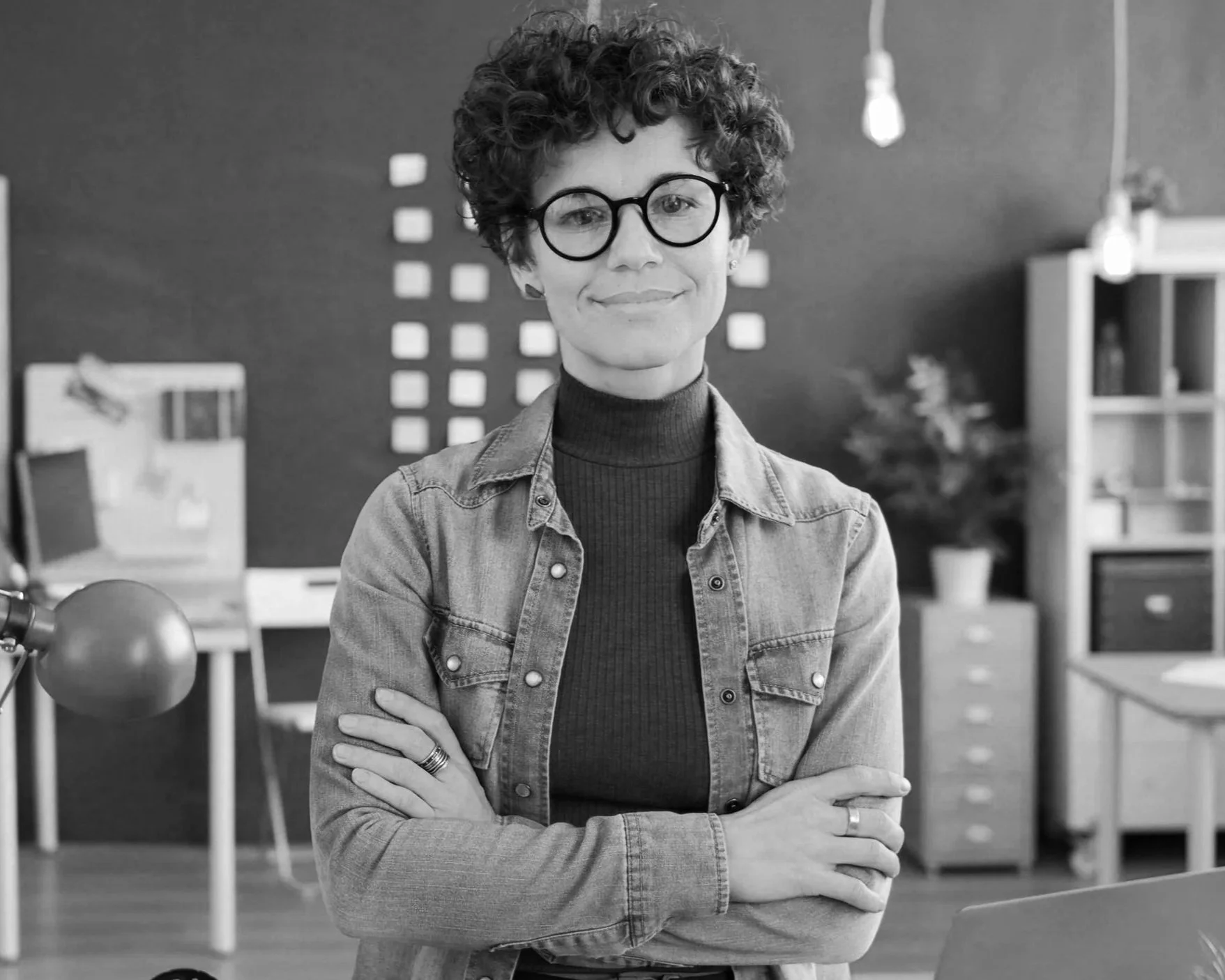 A woman with short curly dark hair and black geometric glasses smiles calmly at the camera. She is wearing a denim shirt over a turtleneck, and her arms are crossed. Behind her is a wall and office furniture.