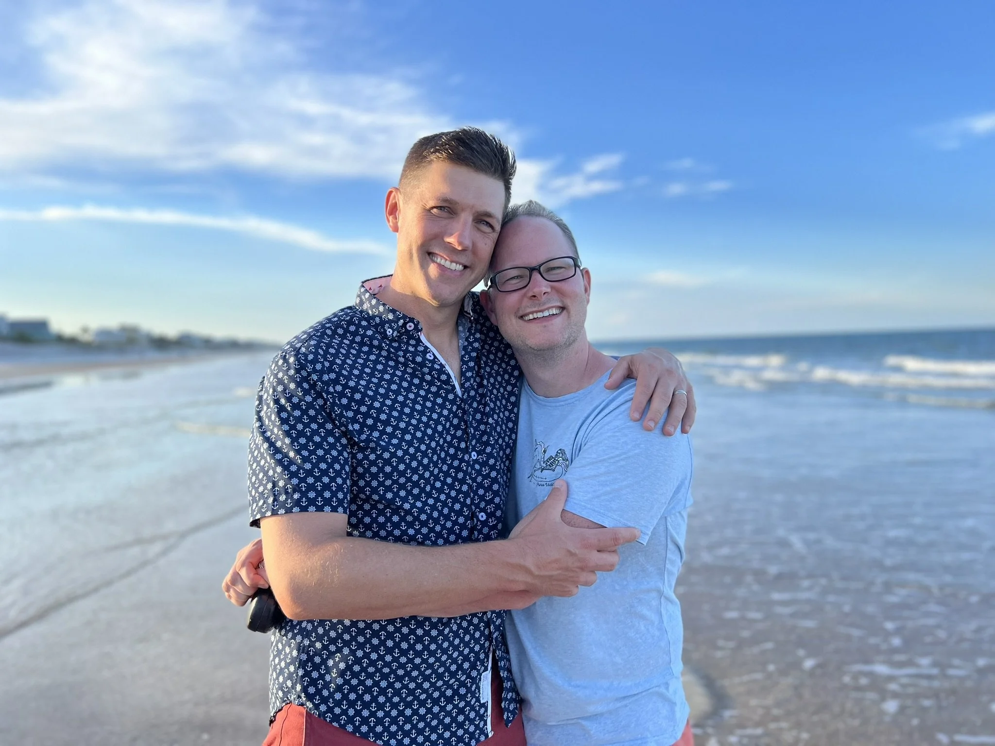 Coach Joe Miller and his husband Matthew stand in a close hug, facing the camera. Joe is wearing a blue shirt with a white pattern, and Matthew is wearing narrow black glasses and a light blue t-shirt. The beach and sky stretch out behind them.