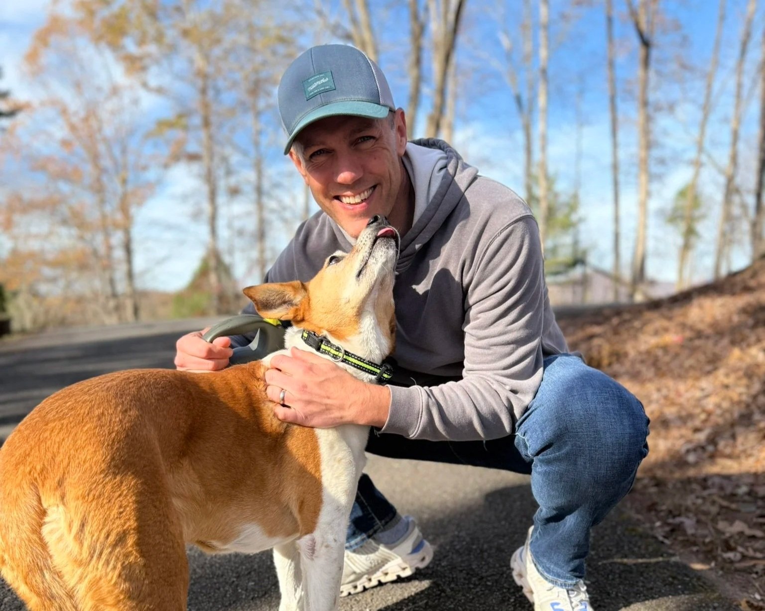 Coach Joe Miller crouches next to a gold-and-white dog. The dog is trying to lick Joe's face, and Joe is grinning at the camera. He's wearing jeans, a gray hoodie, and a blue ballcap. It looks like a crisp fall day with blue skies and fallen leaves.