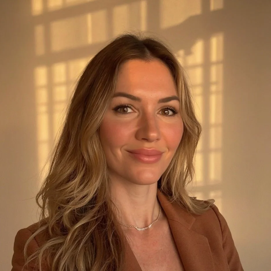 A woman with wavy blonde hair and light skin smiling at the camera. Sunlight creates a grid shadow on the wall behind her.