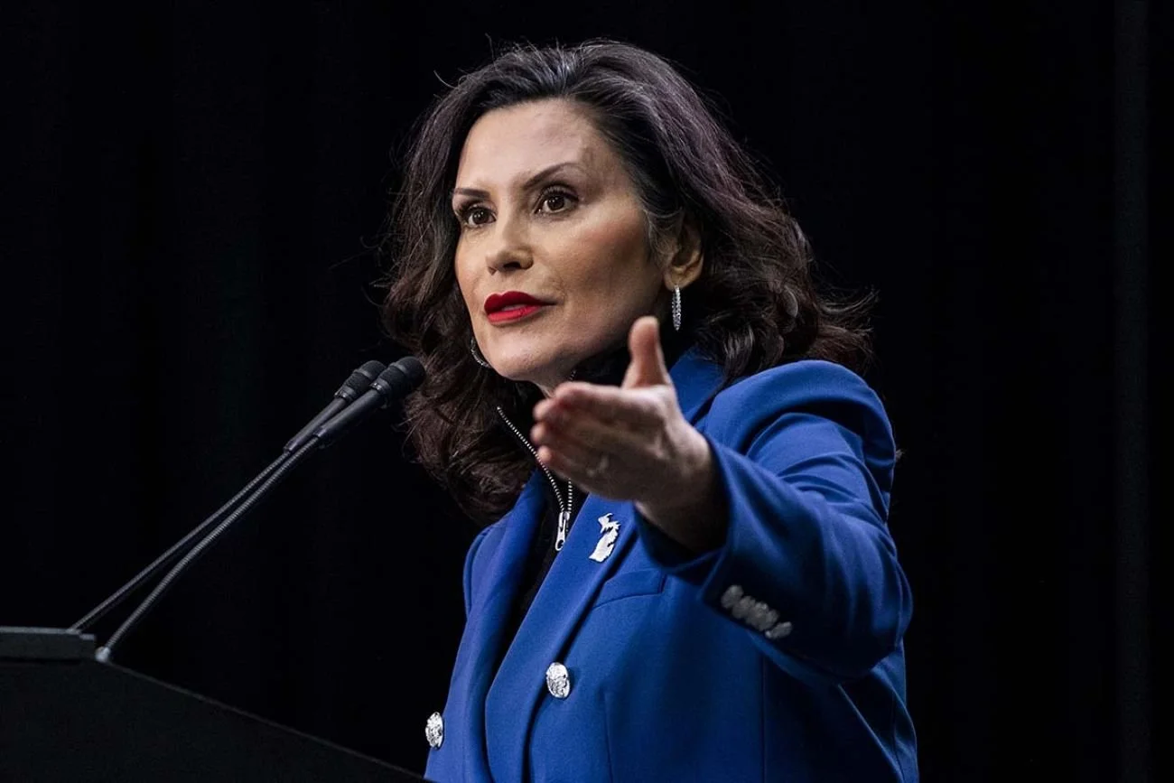 A woman with dark, wavy hair wearing a blue blazer and red lipstick speaking at a podium with a microphone, in front of a dark background.