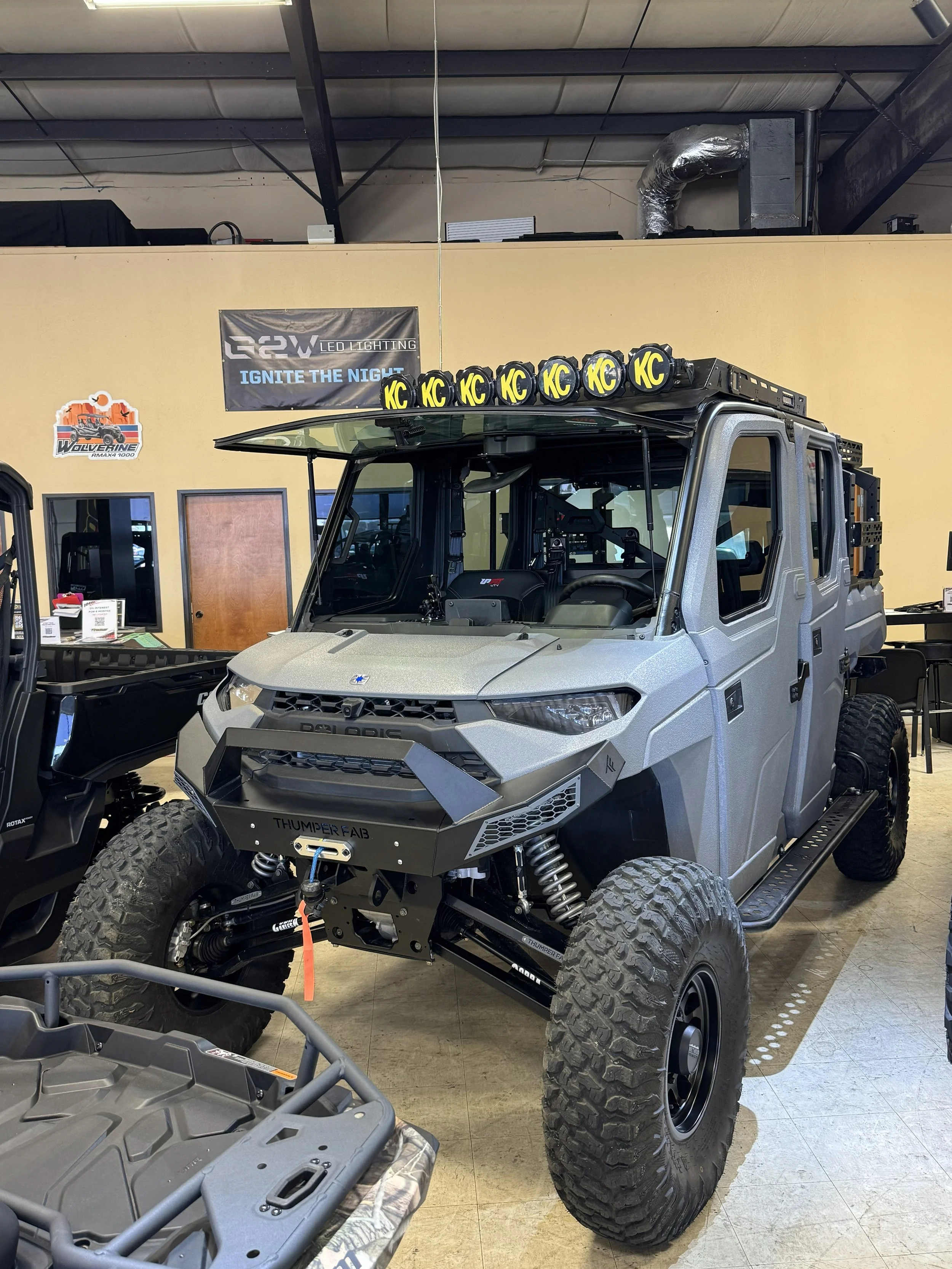 A gray off-road utility vehicle on display indoors, equipped with large tires, a roof-mounted light bar with six KC lights, and custom front and suspension parts.