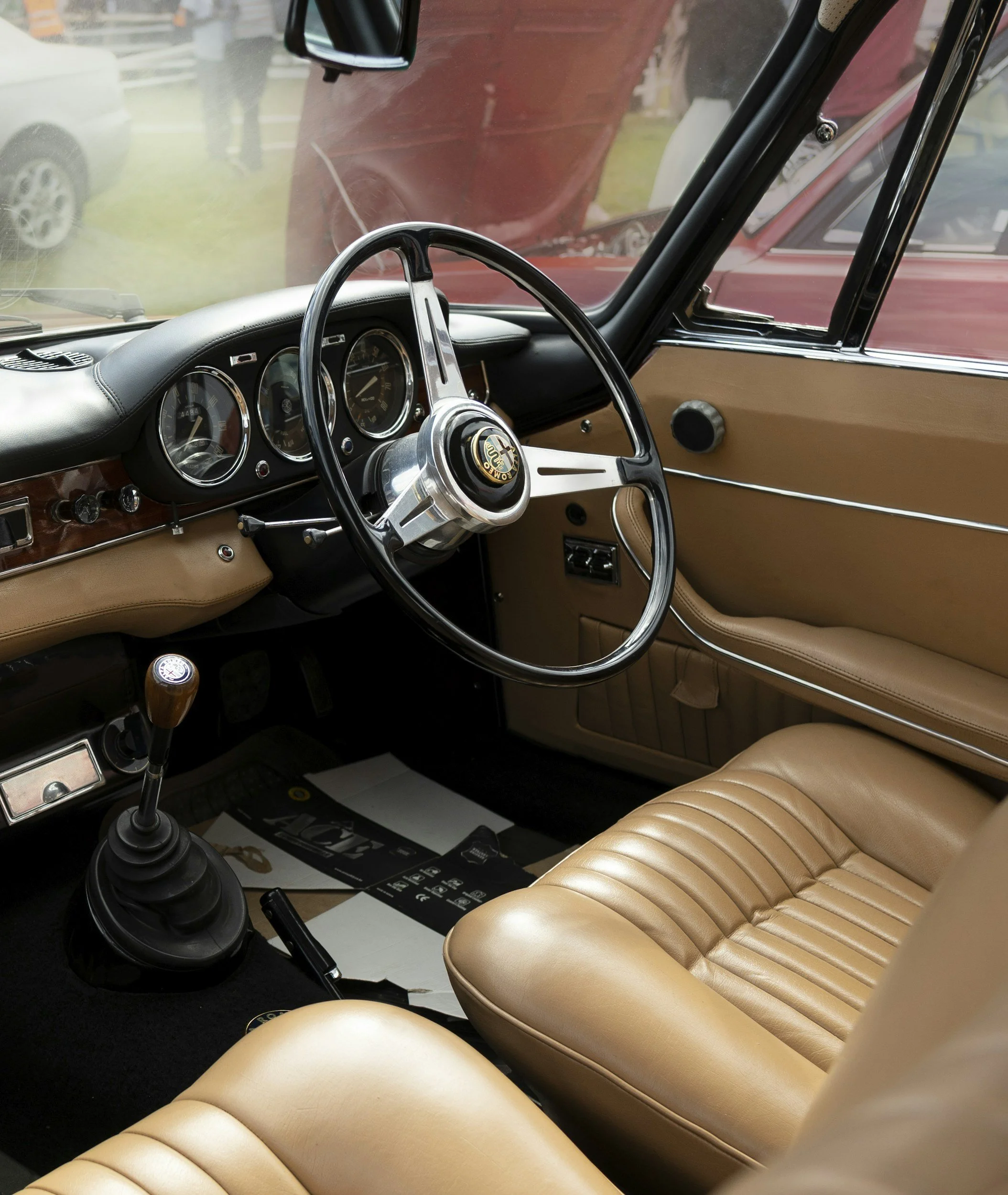 Interior of a vintage car showing a black and silver steering wheel, beige leather seats, dashboard with round gauges, gear shift with wooden knob, and partially visible door with beige interior panels.