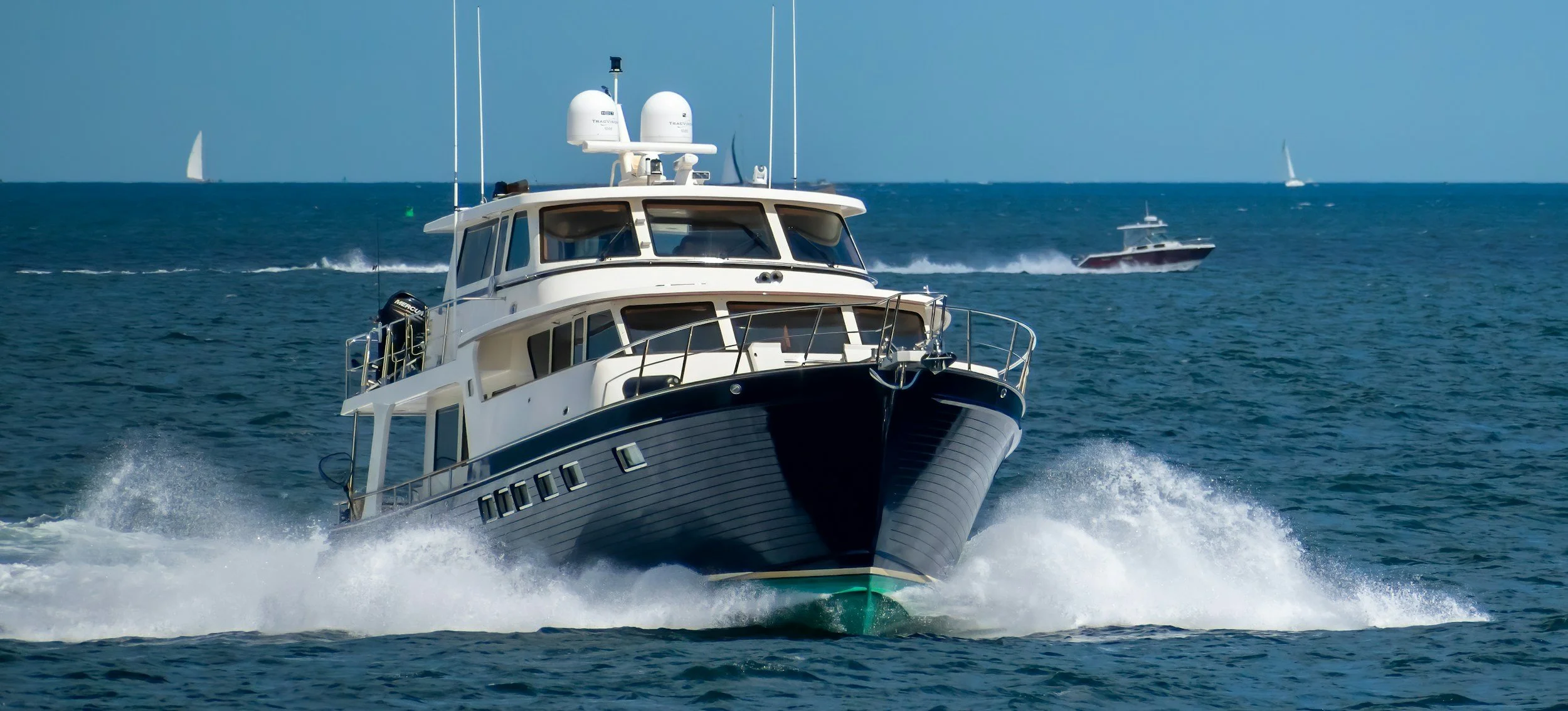 A large white yacht cruising at sea with smaller boats and sailboats in the background.