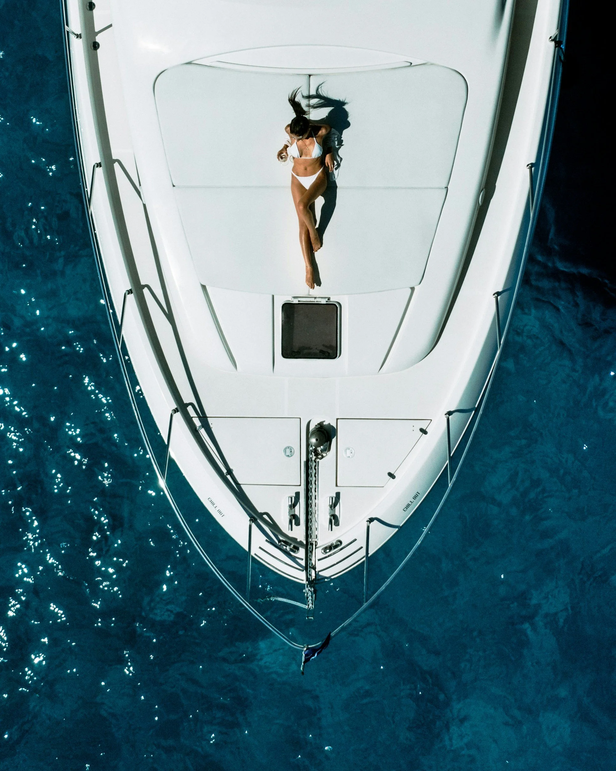 A woman in a bikini lying on the deck of a white yacht, viewed from above, with blue ocean surrounding the yacht.