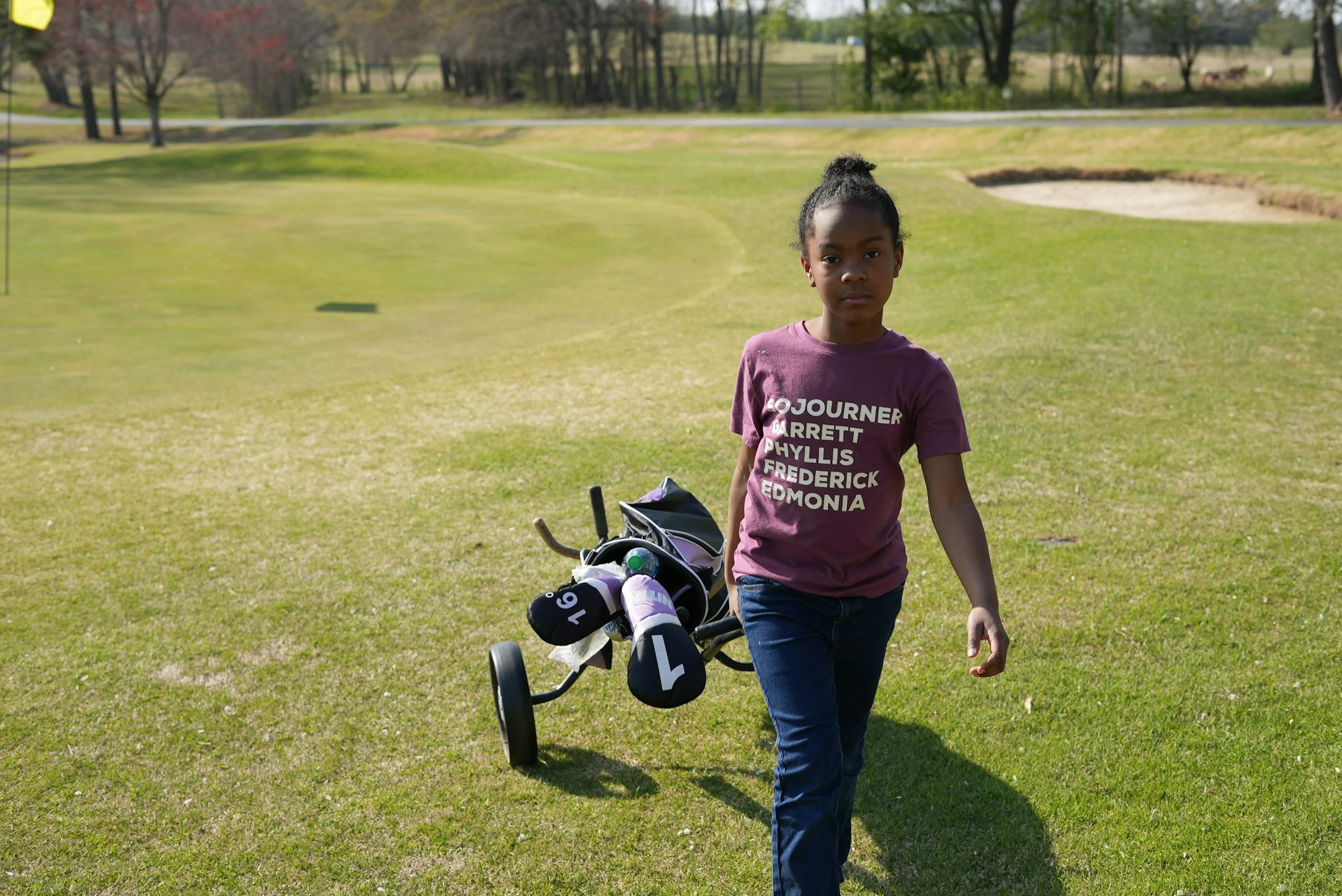 A young girl walking on a golf course holding a golf bag with clubs.