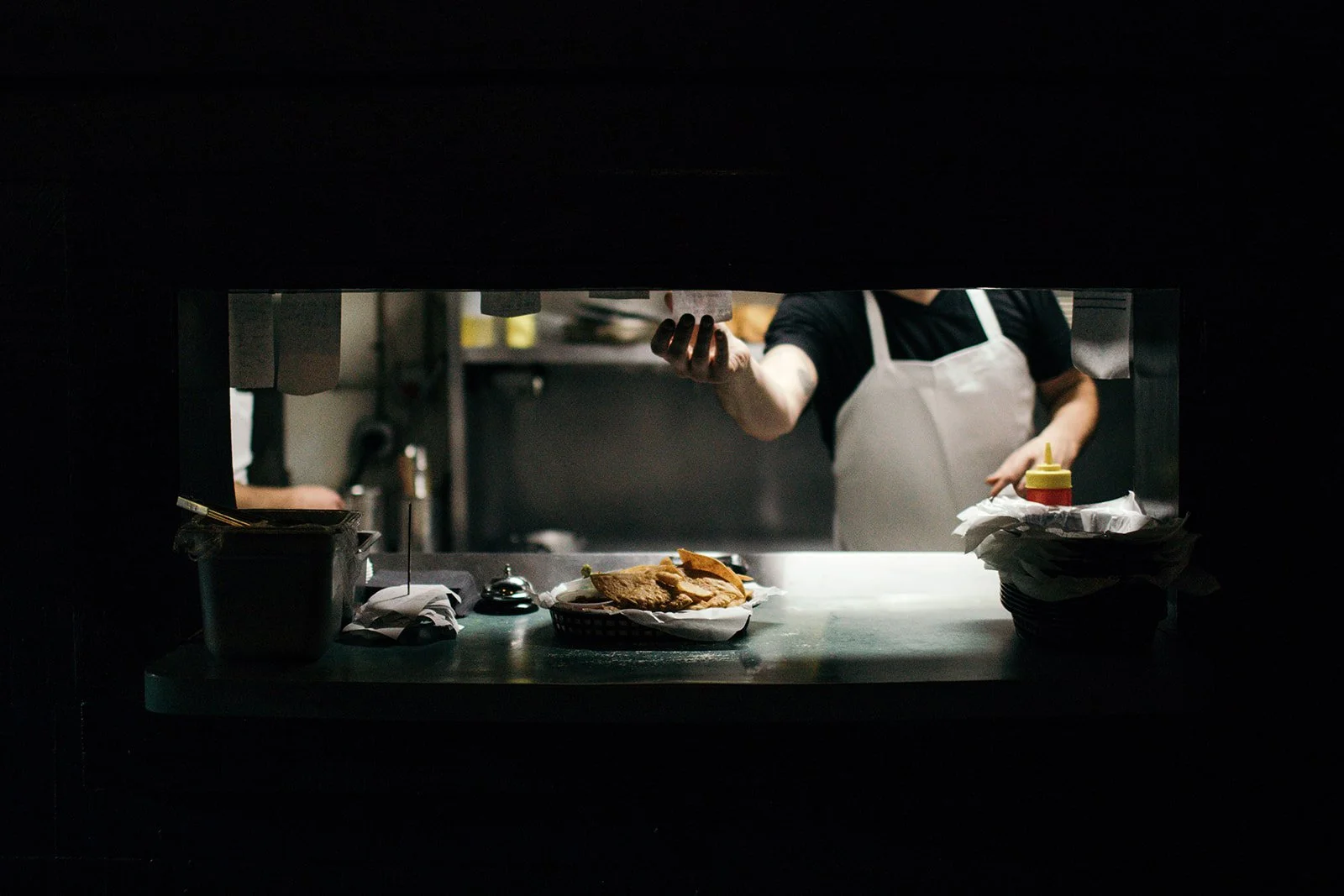 A view through a restaurant kitchen pass, showing a chef in a black shirt and white apron reaching out to receive an order. On the counter below, there are plates with sandwiches, a ketchup bottle, and other kitchen items.