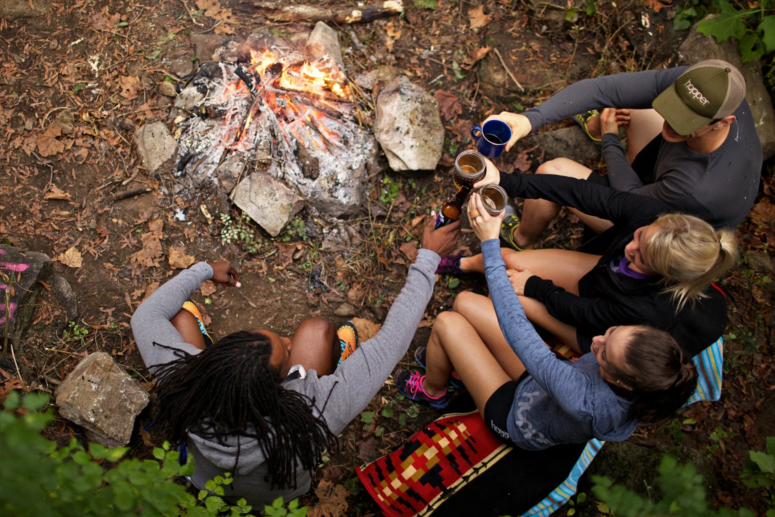 Group of people sitting around a campfire in a wooded area, raising drinks in a toast.