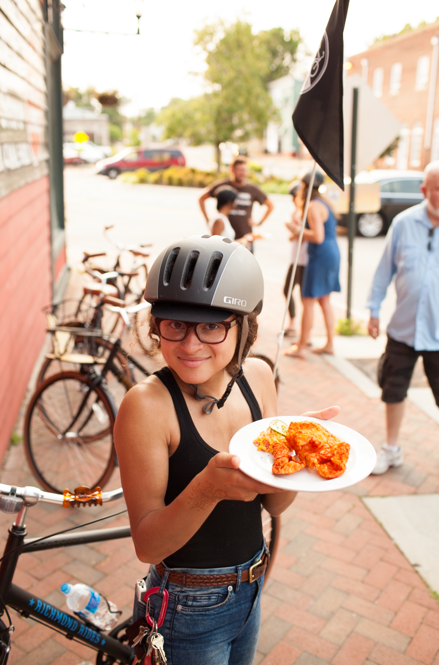 Woman wearing a black helmet and glasses holding a white plate with fried chicken and lemon slices on a city sidewalk, with bicycles, people, and storefronts in the background.