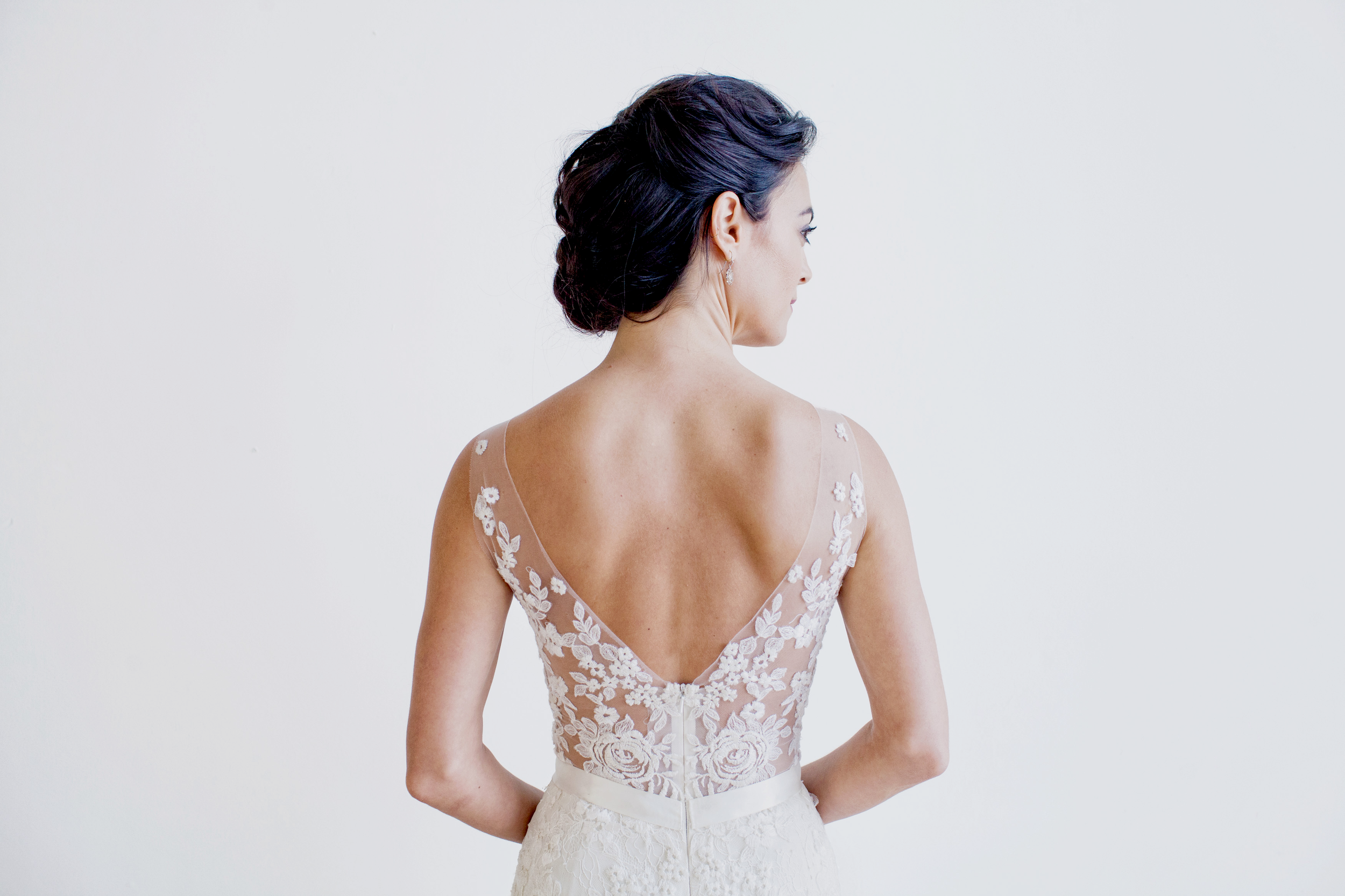 A woman with dark hair styled in an elegant updo, wearing a sheer lace dress with floral embroidery, standing against a plain white background.