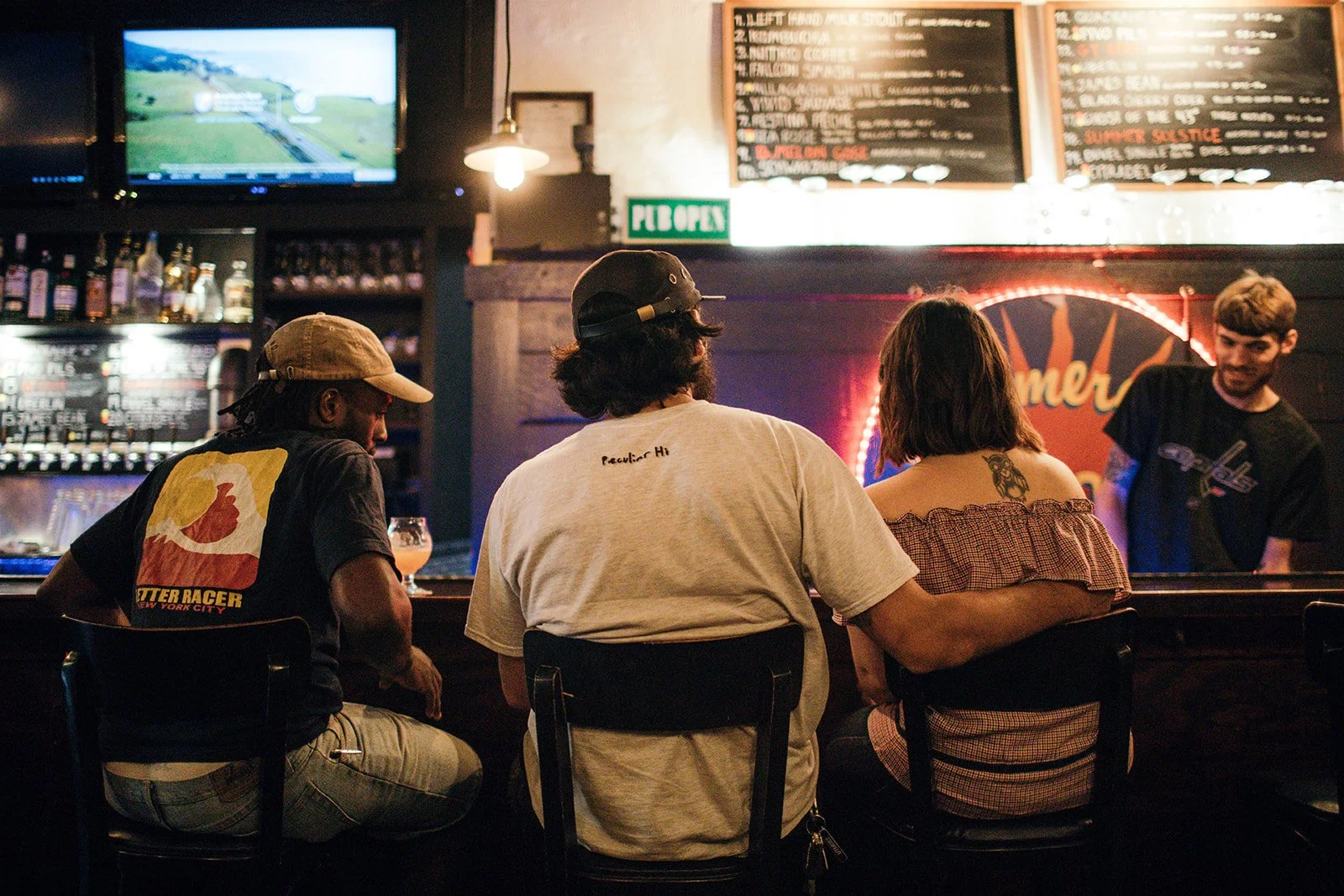 Three friends sitting at a bar counter having a conversation, with a bartender behind the counter. The bar has a sign that says 'FIB OPEN' and chalkboards with menu items above. A TV is mounted in the corner showing a landscape scene. The setting app