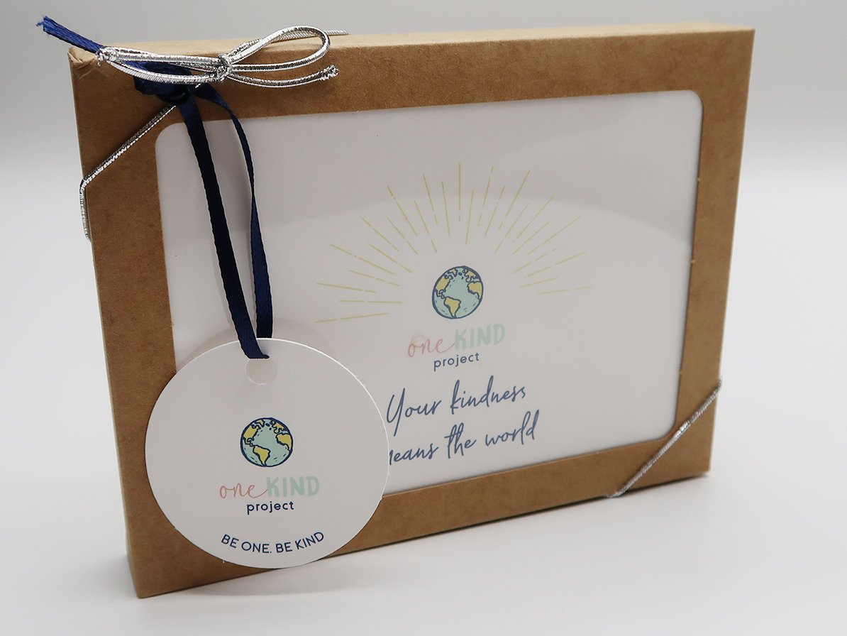 A cardboard box with a clear window displaying a stationery item, adorned with a ribbon and a tag. The tag and box feature a globe illustration and the words "one KIND project" along with the message "Your kindness means the world" and "BE ONE. BE KI