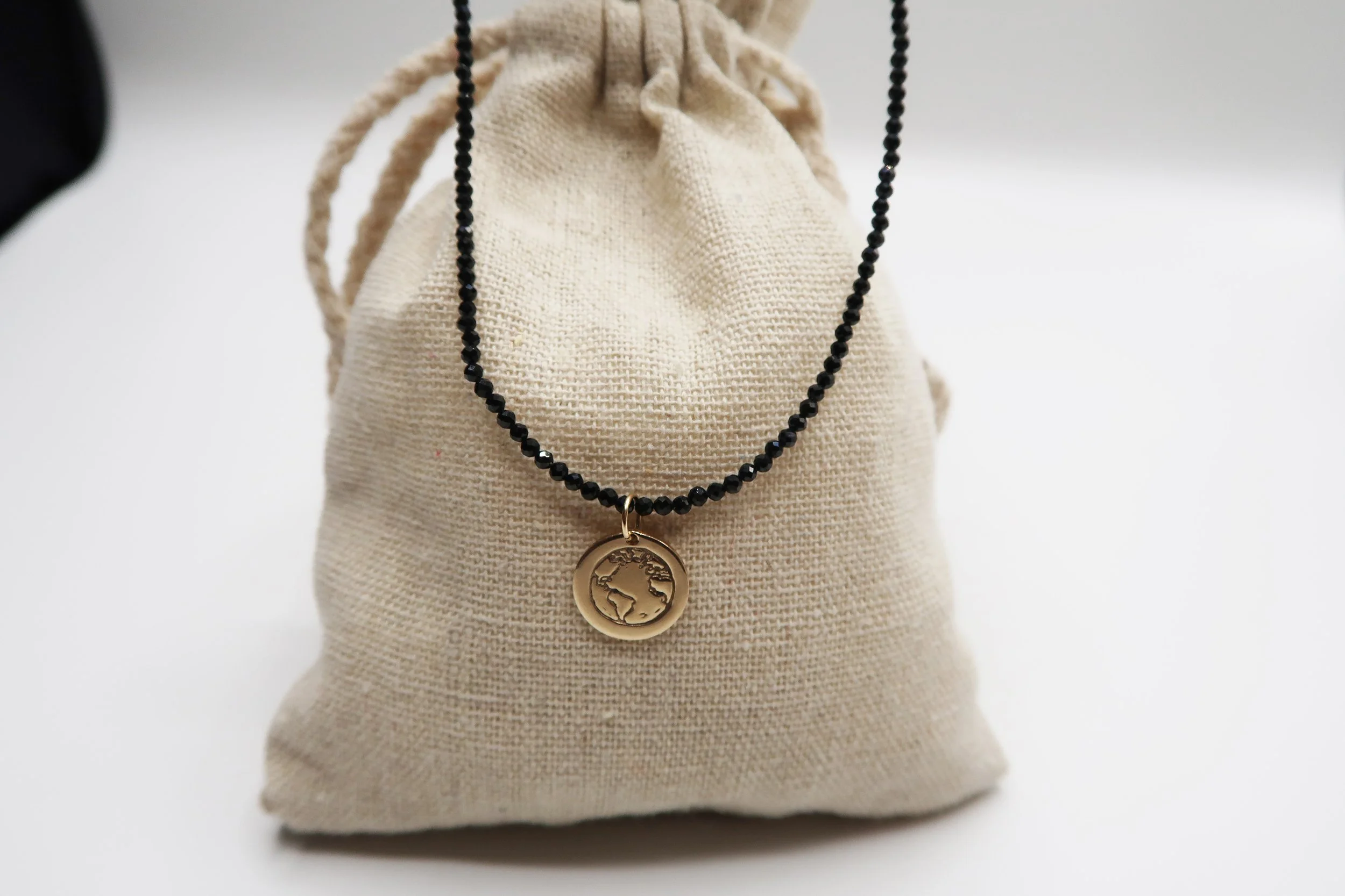 A black beaded necklace with a round gold pendant featuring an engraved map of the world, draped over a beige fabric pouch.