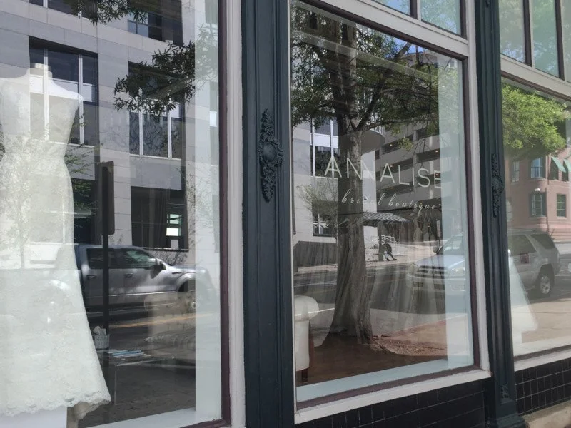 Storefront window display with a large tree and city street reflected in the glass, showing parked cars and buildings.