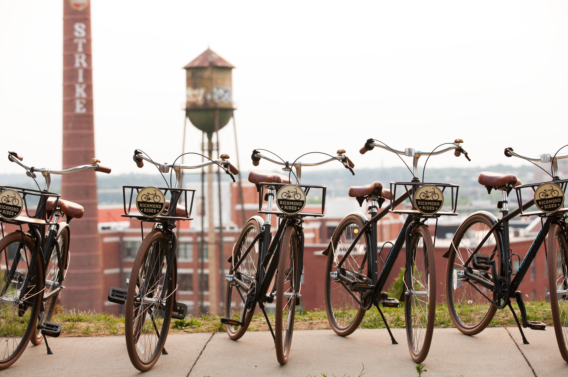 A row of bicycles parked outdoors with a brick building and a water tower in the background.