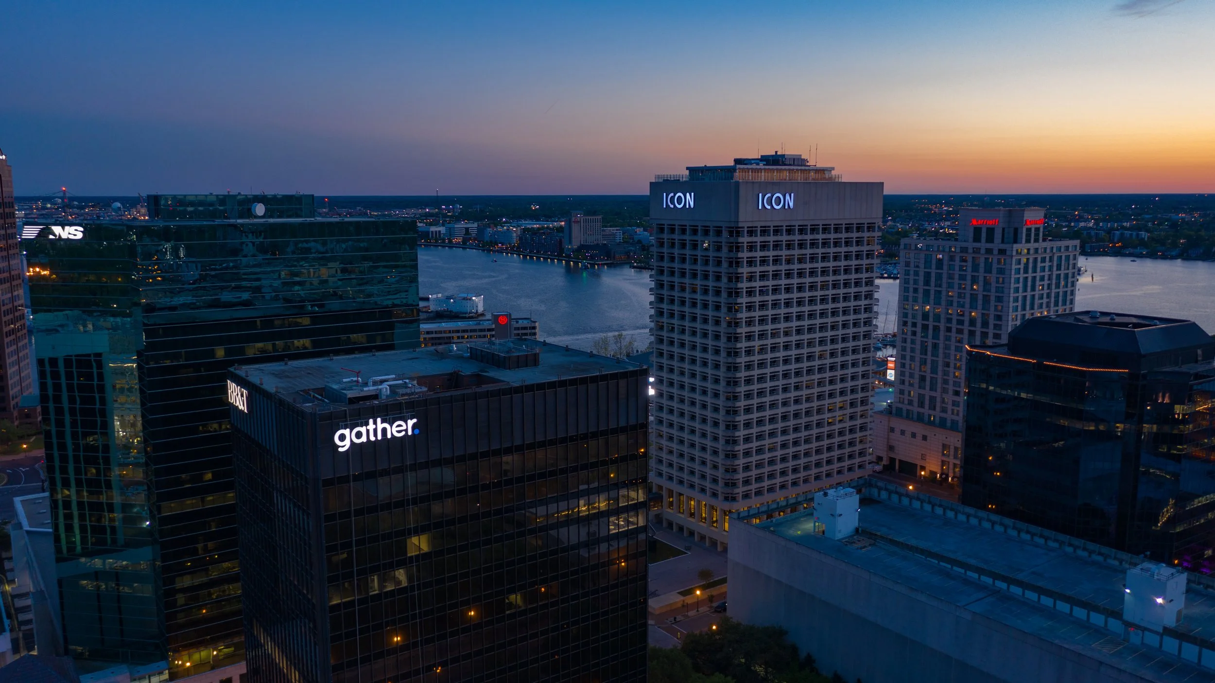 Skyline view of a city at dusk with tall buildings, illuminated signs including 'gather' and 'ICON,' and a river running through the city.