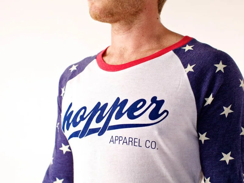 Close-up of a man wearing a white and blue baseball-style shirt with red trim, featuring the words 'hopper APPAREL CO.' on the front, and stars on the sleeves.