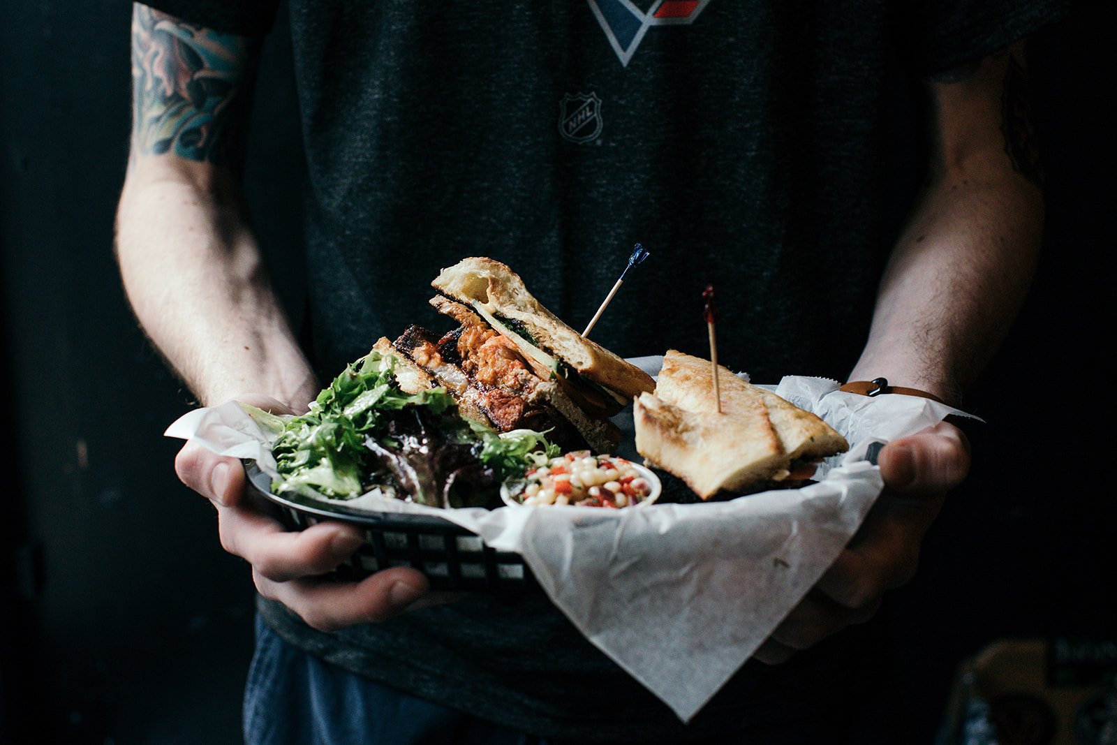 Person holding a black basket with a sandwich, salad, and side, lined with white paper.