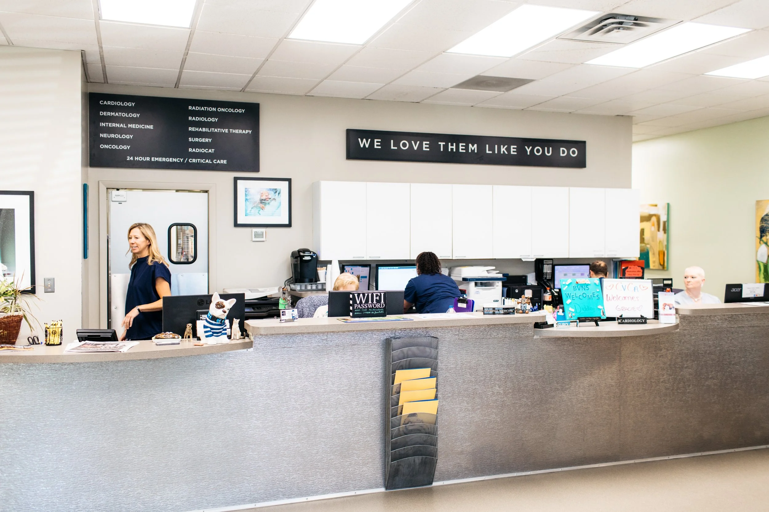 Hospital reception area with staff working at computers, a woman standing and smiling, and signs indicating medical departments.