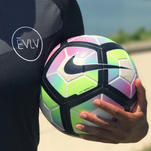 Close-up of a person holding a colorful Nike soccer ball outdoors with water and greenery in the background.