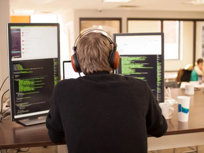 Person wearing headphones working on multiple computer screens with code in a modern office