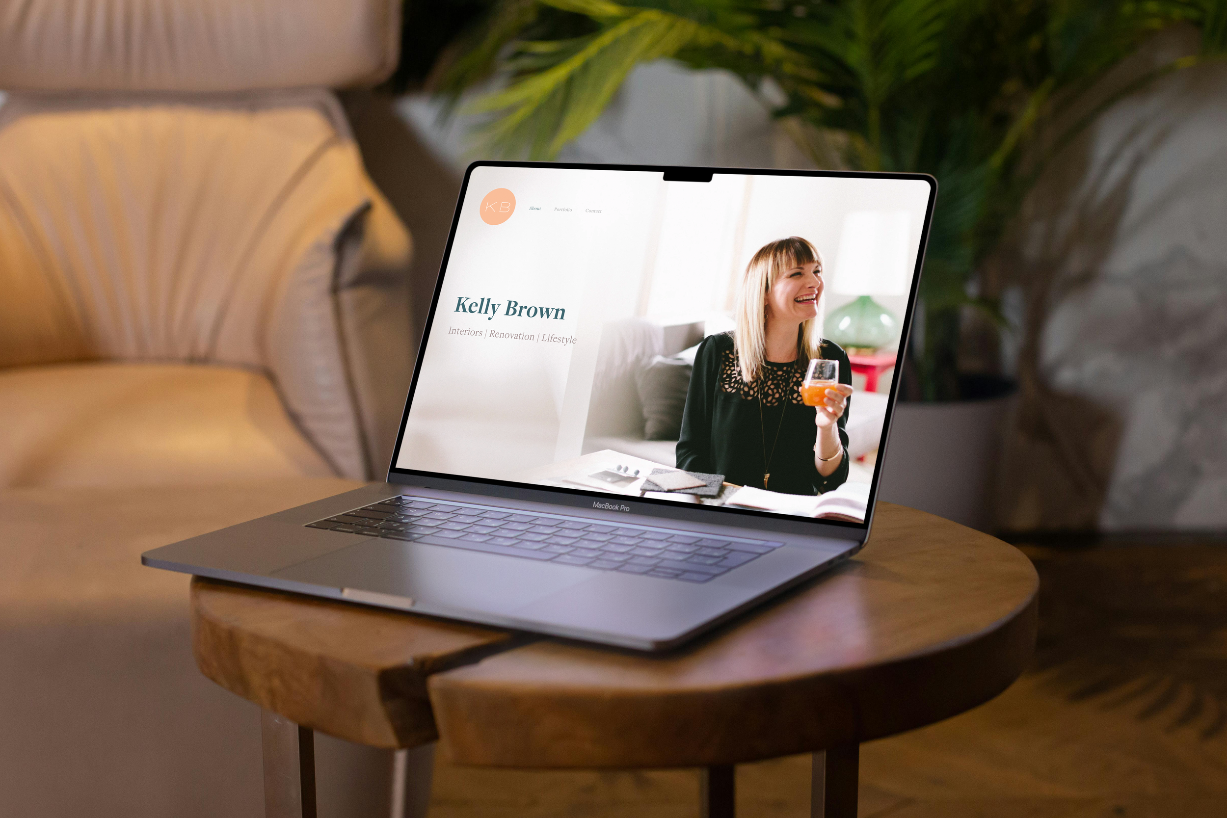 A laptop on a wooden table displays a website featuring a woman smiling and holding a glass, with the text 'Kelly Brown' and links to 'About,' 'Portfolio,' and 'Contact.'