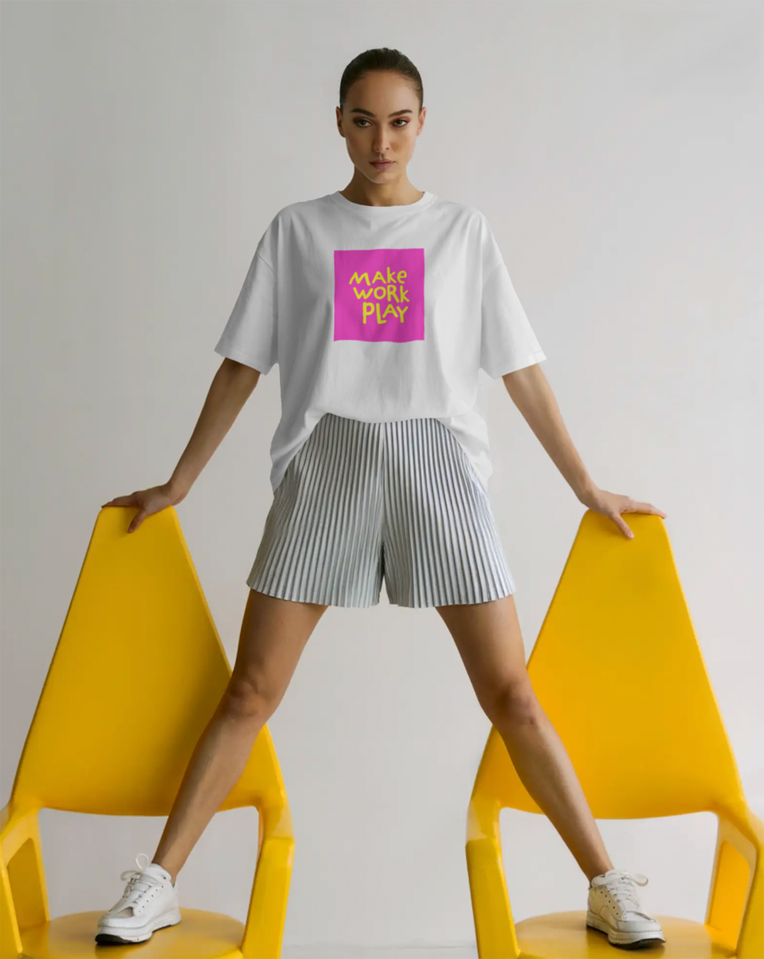 A young woman standing between two yellow chairs, holding the edges of the chairs with her hands, wearing a white t-shirt with a pink square and neon green text that says 'MAKE WORK PLAY,' striped shorts, and white sneakers, against a plain white bac