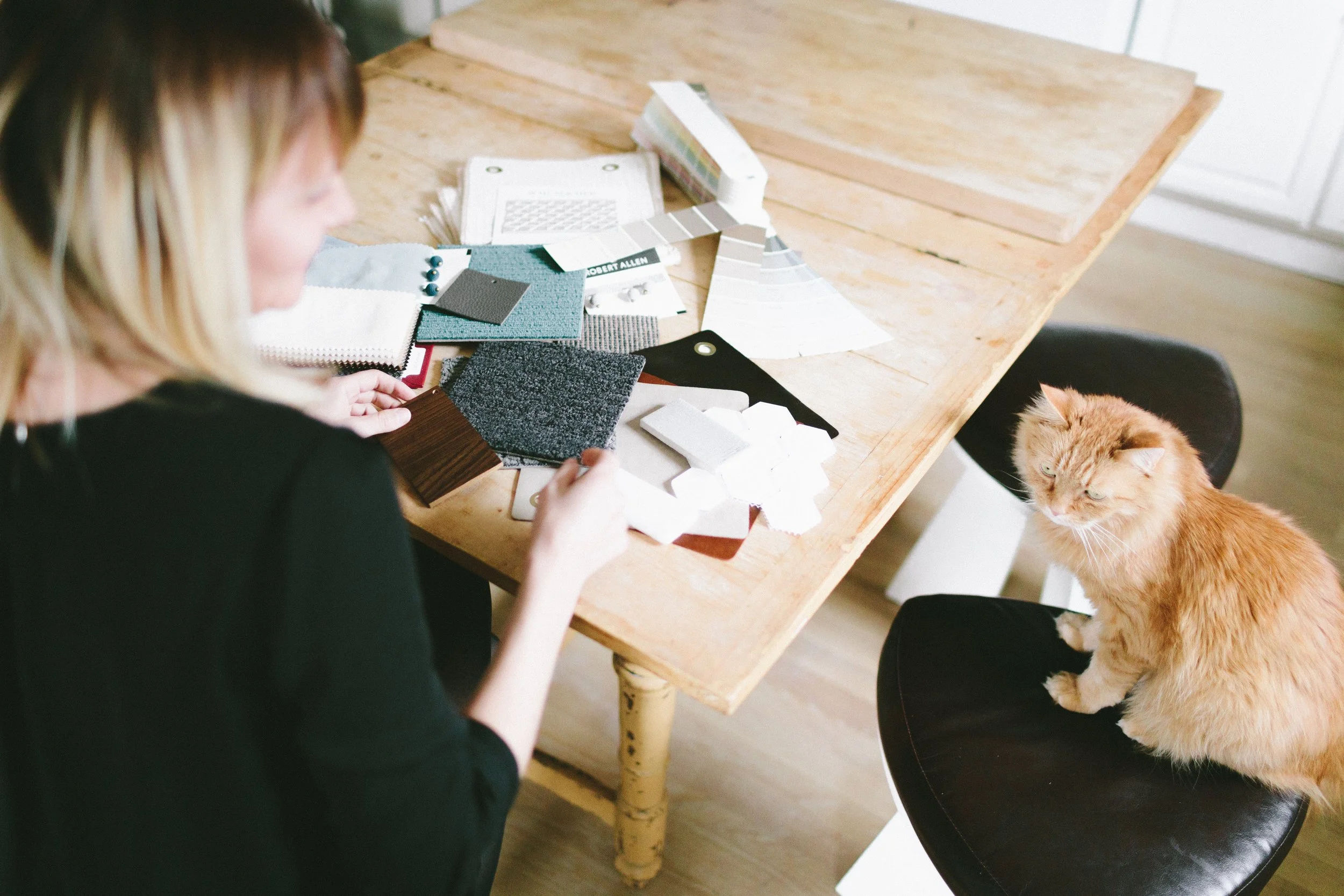 A woman with blonde hair arranging fabric samples on a wooden table with a ginger cat sitting nearby on a black chair.