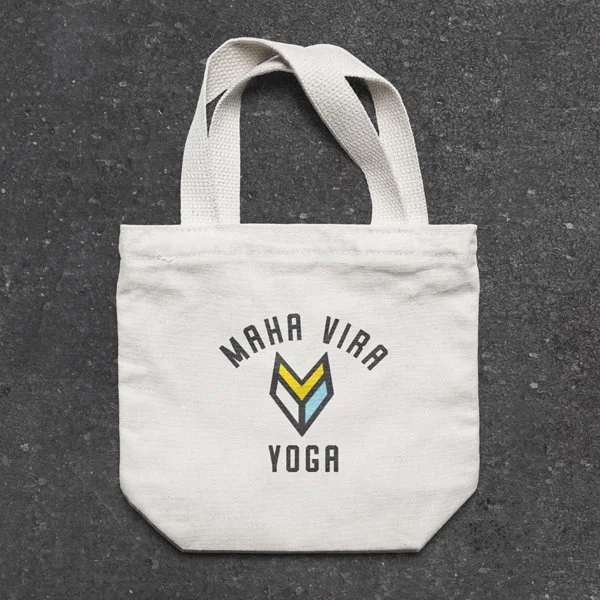 A white tote bag with the text 'Maha Vira Yoga' and a geometric logo printed on it, lying on a dark textured surface.